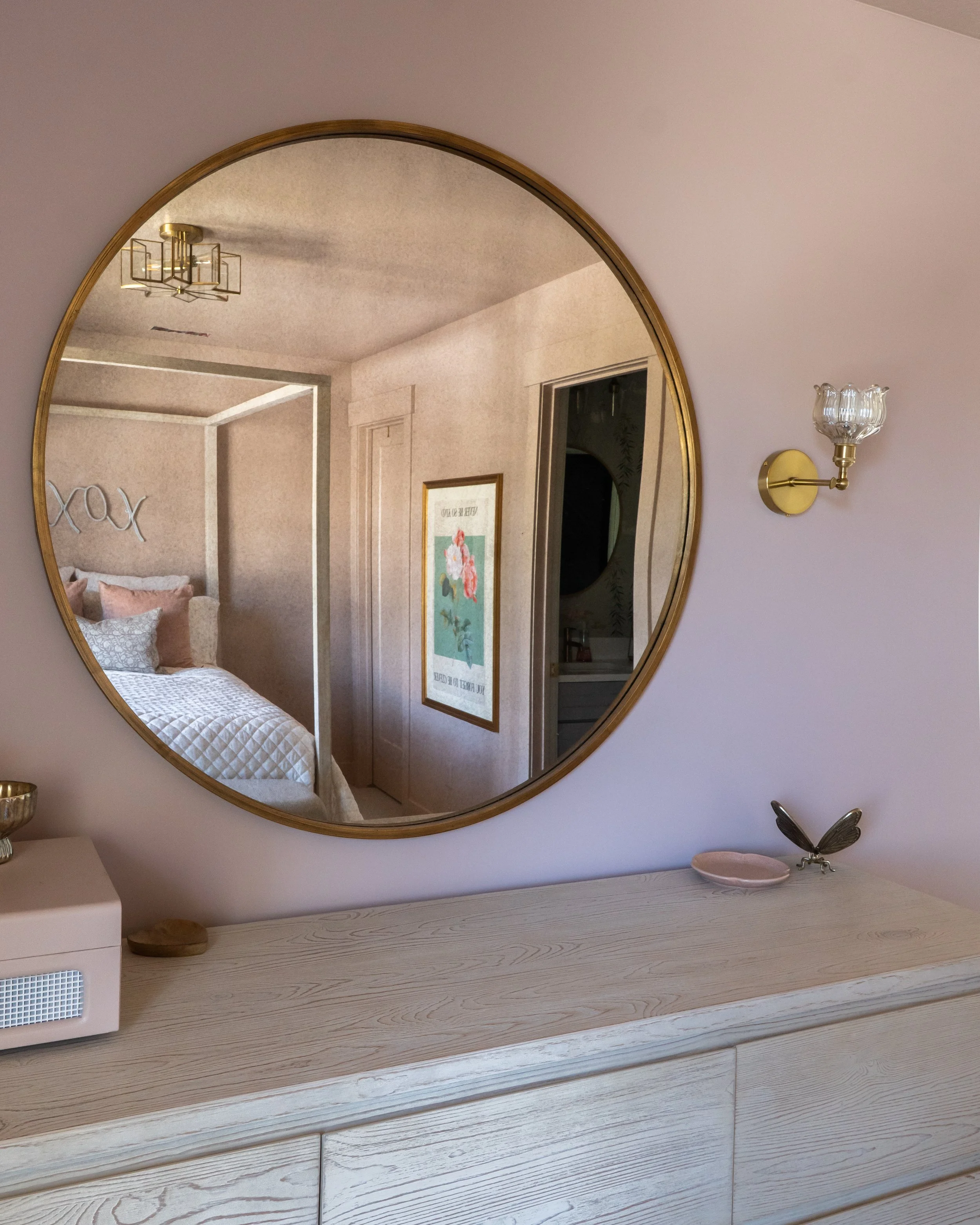 A round mirror on a light-colored wooden dresser reflects part of a bedroom with a hanging light fixture, a bed with white quilt, pillows, a framed artwork, and a doorway. A decorative lamp with a glass shade and a butterfly figurine are on the dress