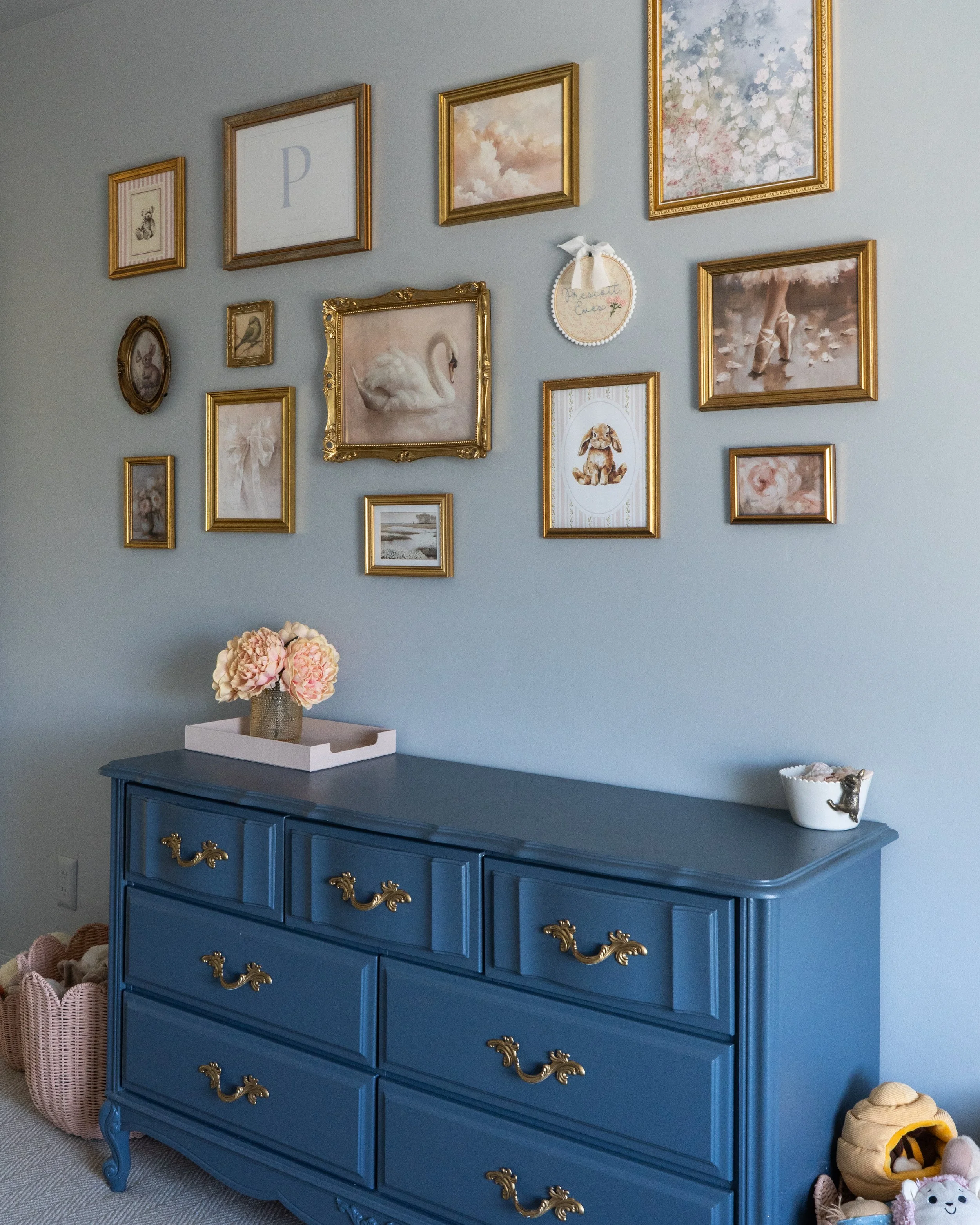 A blue vintage dresser with gold handles and accents against a light blue wall decorated with various framed pictures including animals, clouds, and a ballet dancer. A white tray with pink peonies sits on top, and baskets with stuffed animals are on 