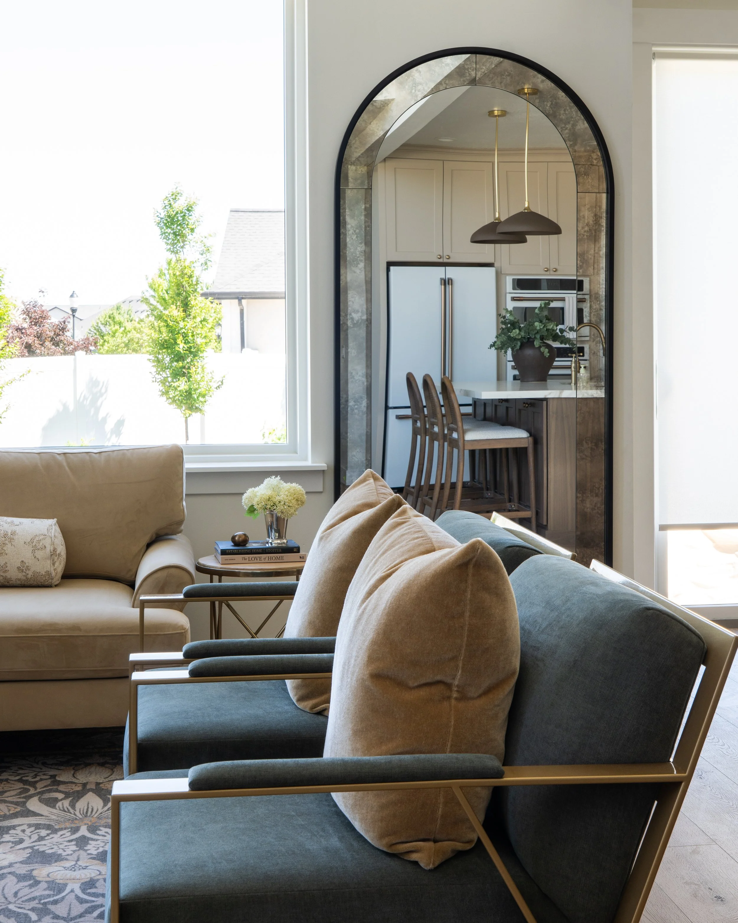 A bright living room with beige sofa and a side table with flowers, with a large window showing green trees outside. In the background, beyond a decorative archway, kitchen area with white cabinets, a refrigerator, and pendant lighting. The room has 