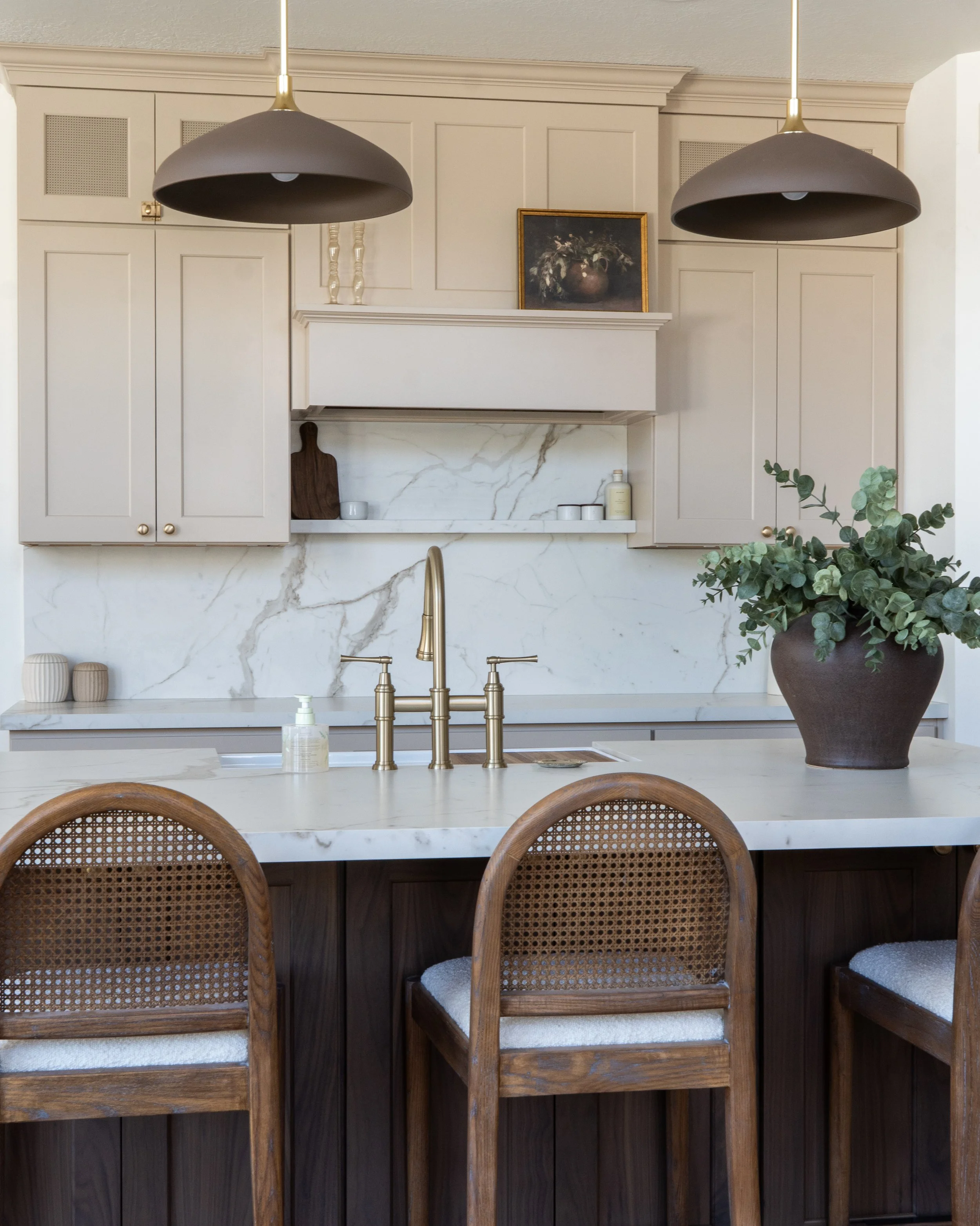 Kitchen with beige cabinetry, marble backsplash, gold fixtures, two pendant lights, potted plant, and wooden chairs at the island.