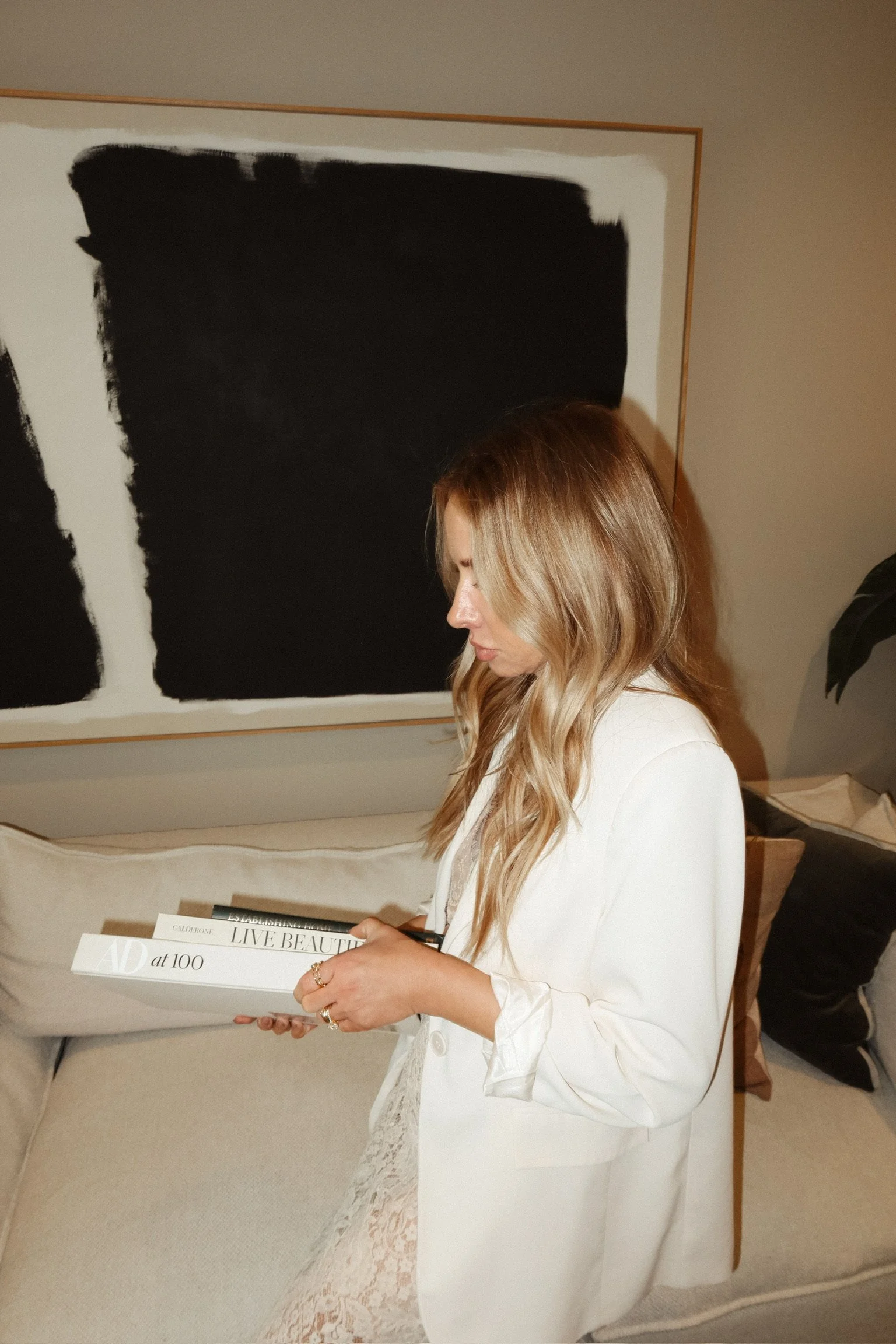 A woman with long wavy blonde hair looking down at a magazine or booklet in her hands, wearing a white blazer and sitting on a light-colored bed or sofa with a large black and white abstract painting on the wall behind her.