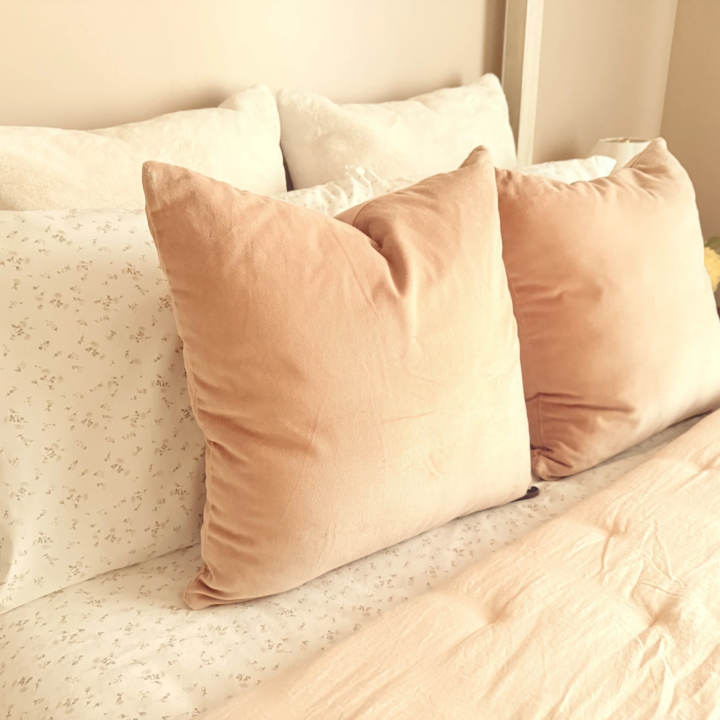 Close-up of a bed with multiple pillows, including light beige and tan velvet pillows, in a bedroom with a beige wall and floral-patterned bedsheet.