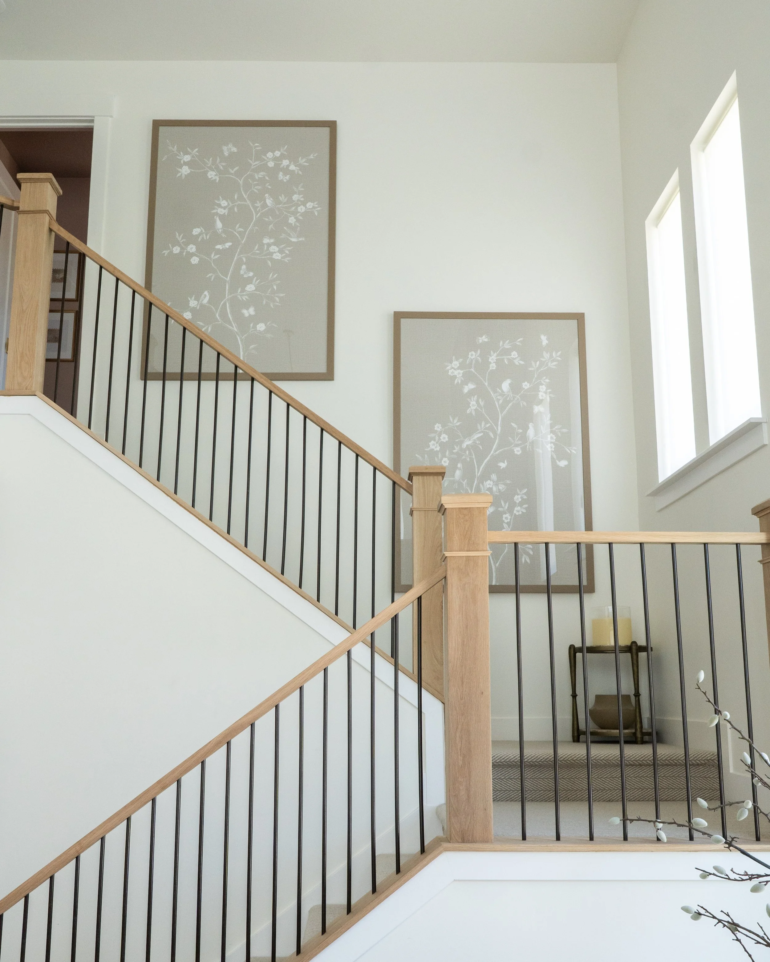 Upper view of a modern staircase and landing with light-colored wood and black metal railing, along with two large framed art pieces of white tree branches with birds on a beige background on the white wall. Sunlight streams through two windows on th
