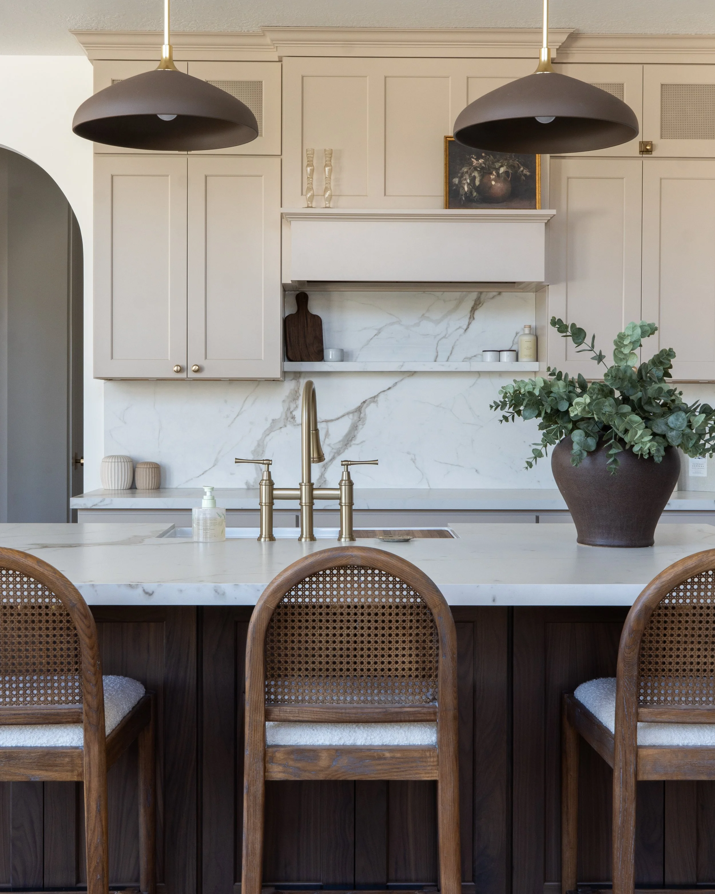 A kitchen scene featuring beige cabinets, a marble backsplash, a large pot with greenery, and a white countertop with three wooden chairs in front.