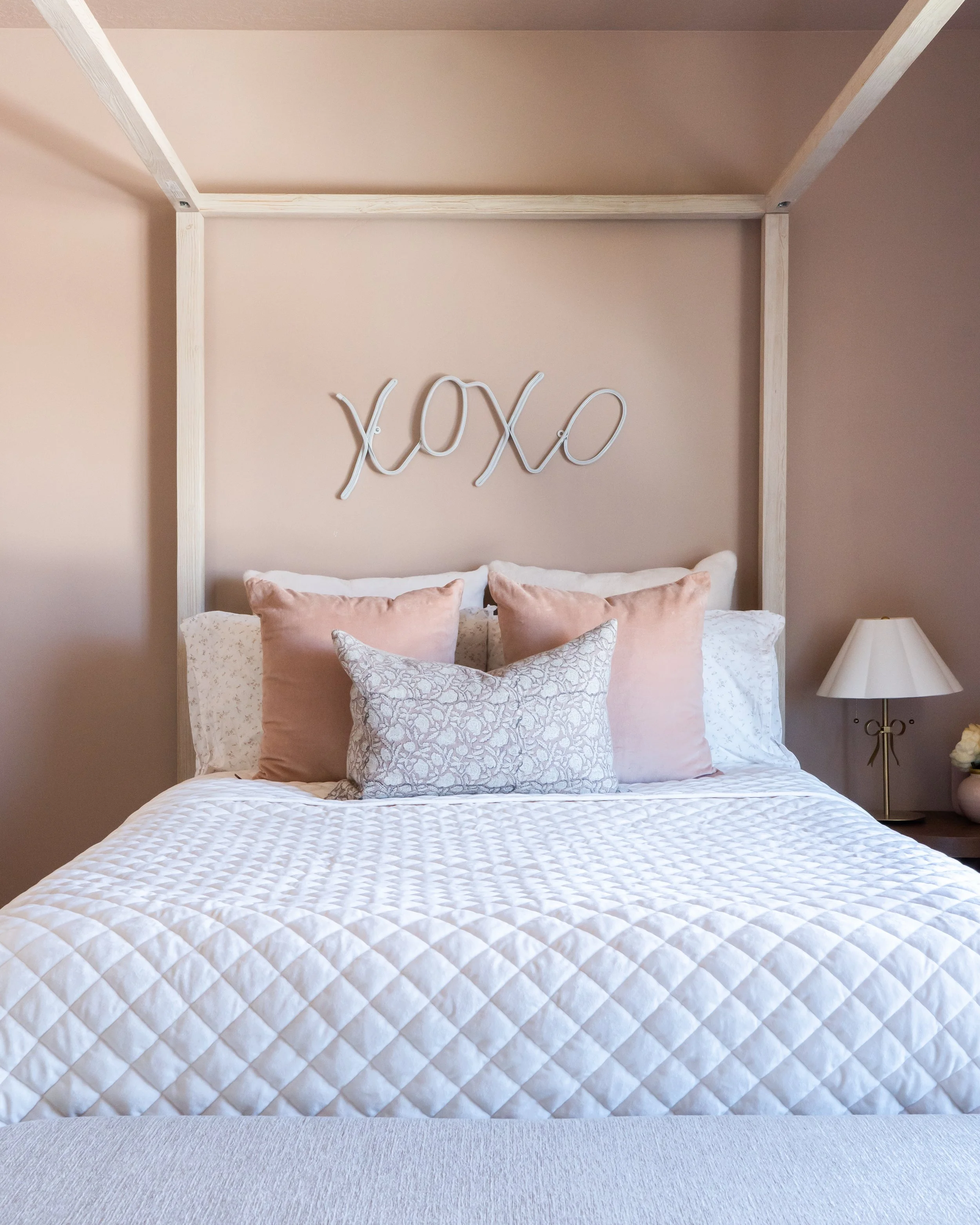 A modern bedroom with a bed featuring a white quilted bedspread and multiple pink and patterned pillows. Above the bed, there is a simple decorative sign spelling 'XOXO' in neon lights. To the right, a white bedside lamp with a beige shade sits on a 