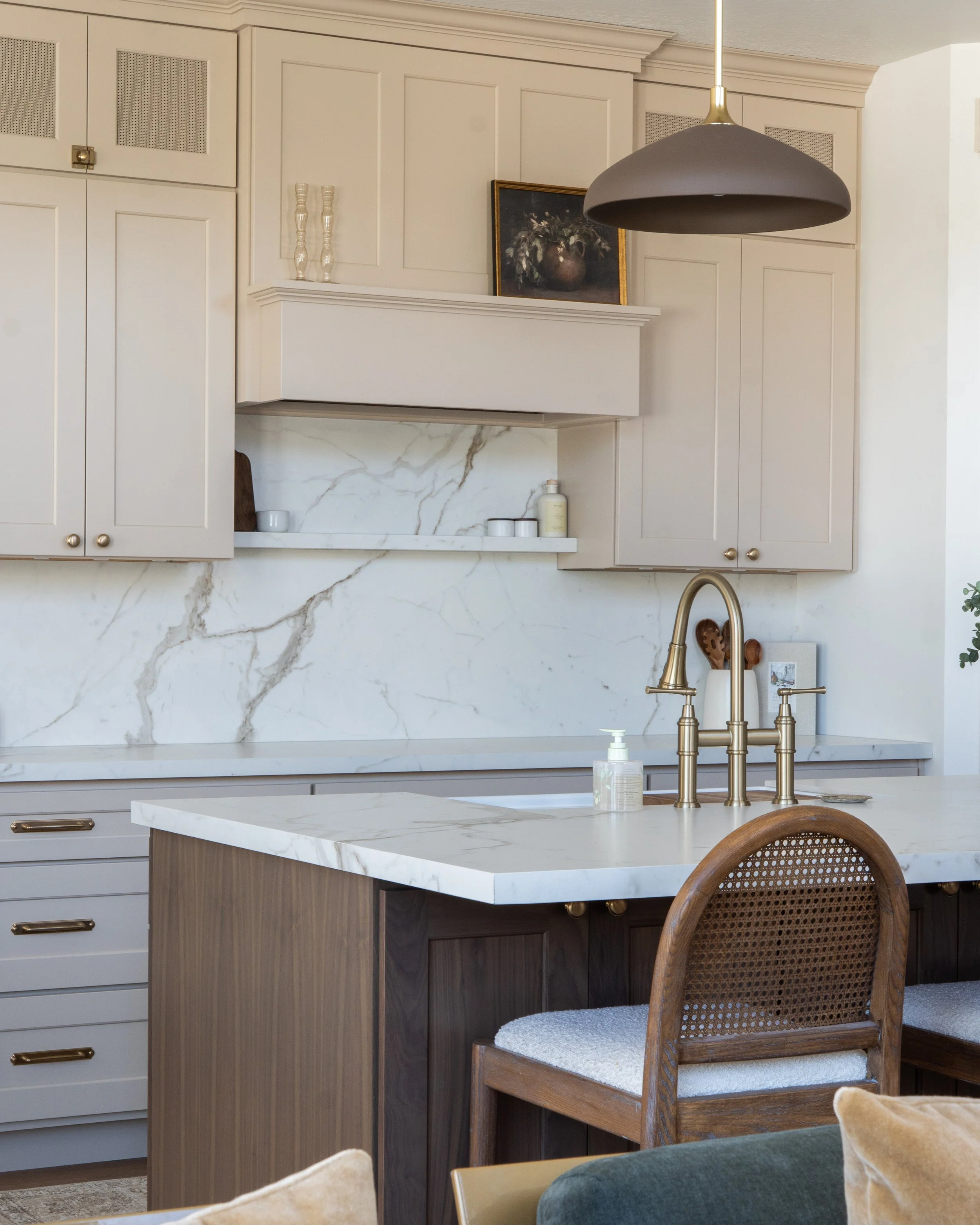 A modern kitchen with a white marble countertop and backsplash, beige and dark wood cabinets, a gold faucet, and decorative items including a framed painting and glass vases.