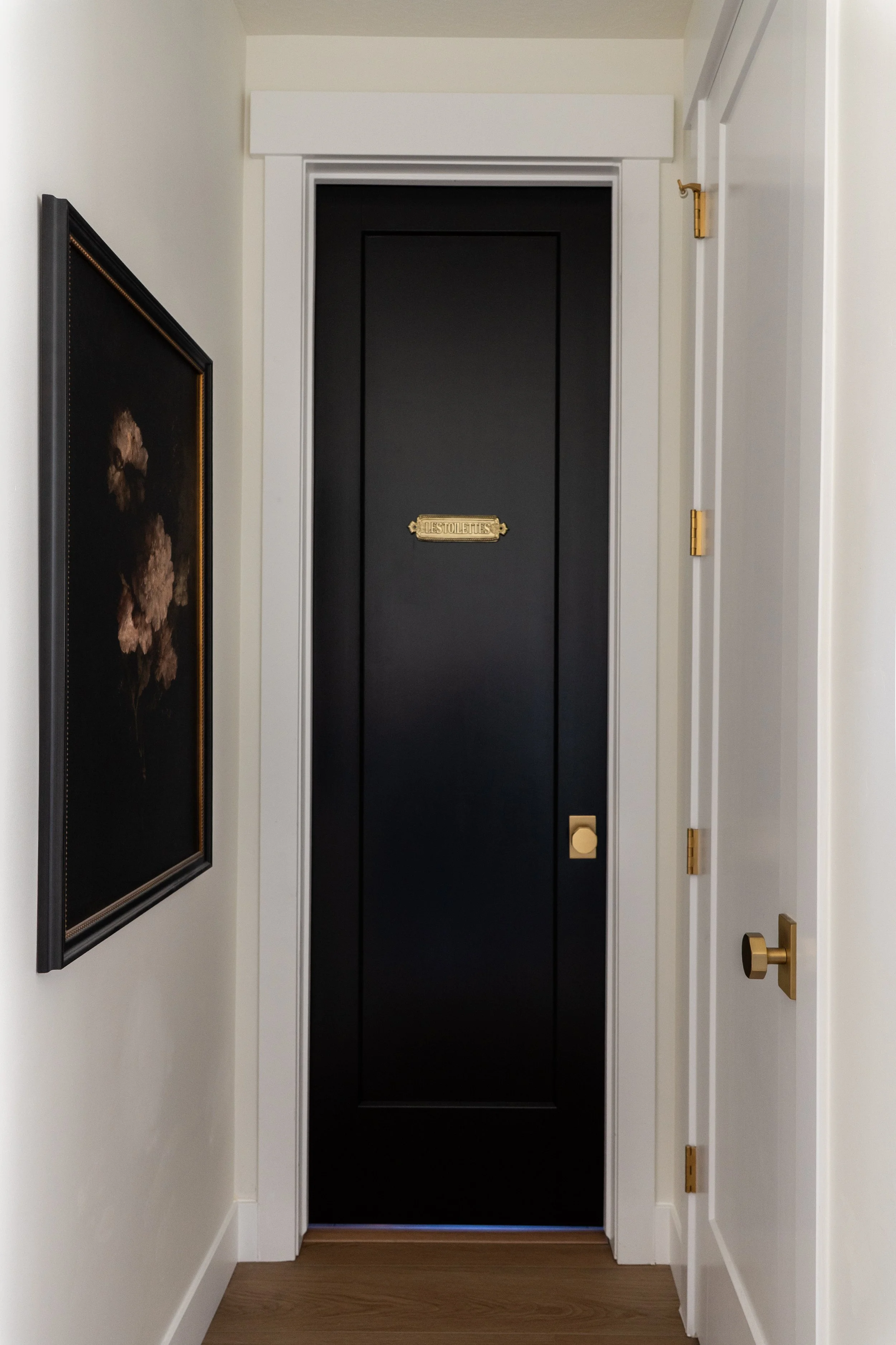 A hallway with white walls and a black door labeled "Les Toilettes" in gold. The hallway has a framed painting on the left wall and gold hardware, including hinges and a door handle, on the right door.