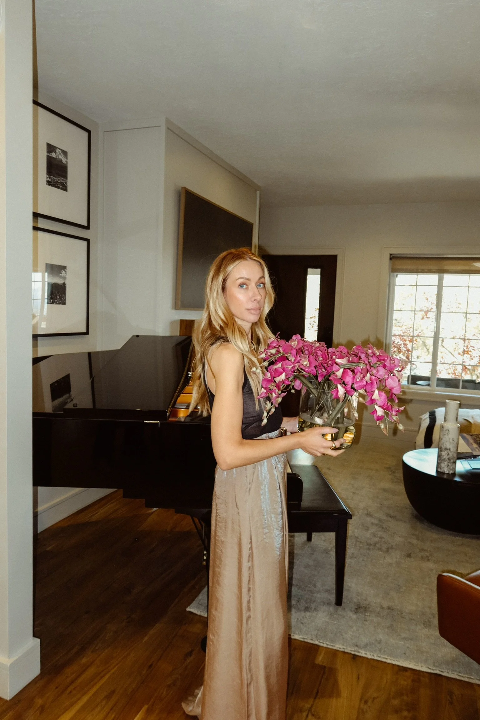 A woman with long blonde hair holding a bouquet of pink flowers in a living room with a black grand piano, framed black-and-white photographs on the wall, a window with trees outside, and modern furniture.