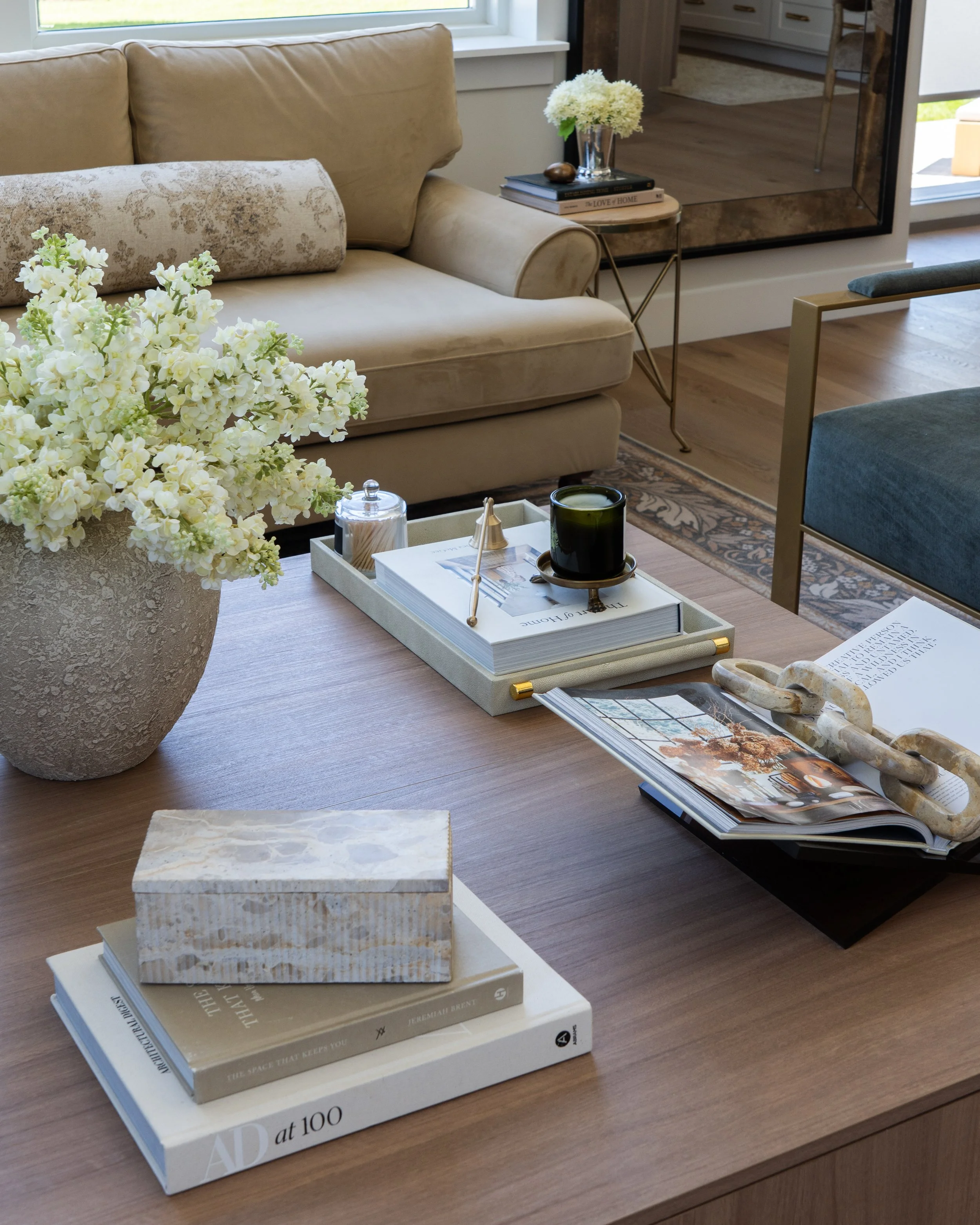 Living room with beige sofa, side table with flowers and books, coffee table with open magazines, decorative sculptures, and a window in the background.