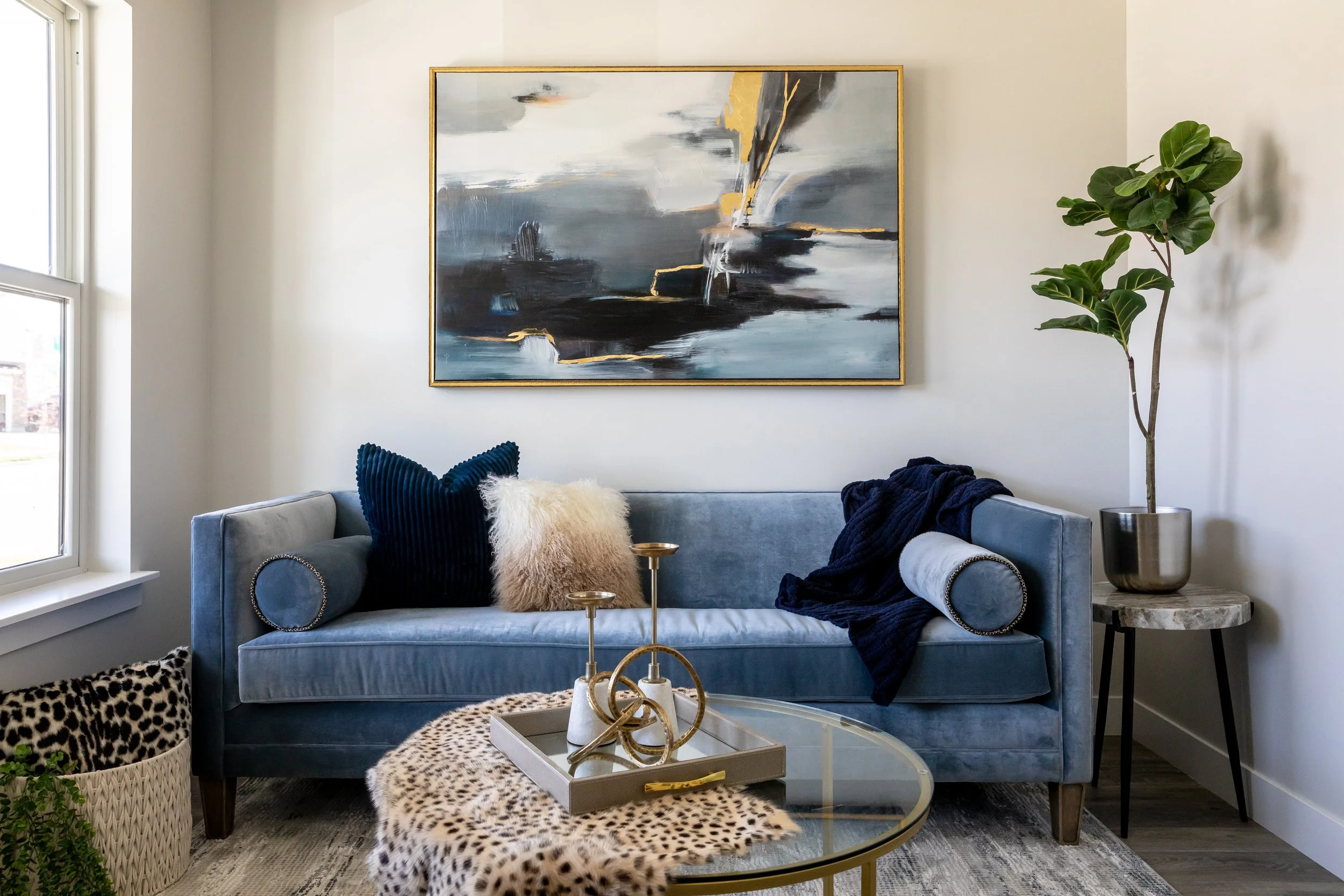 A living room with a blue velvet sofa decorated with a fluffy cream pillow and a navy blue pillow, a glass coffee table with golden decorative items, a leopard print cloth, a painting with abstract gray, black, and gold colors hanging on a white wall, a large potted fiddle leaf fig plant, and a marbled side table with a metallic planter.