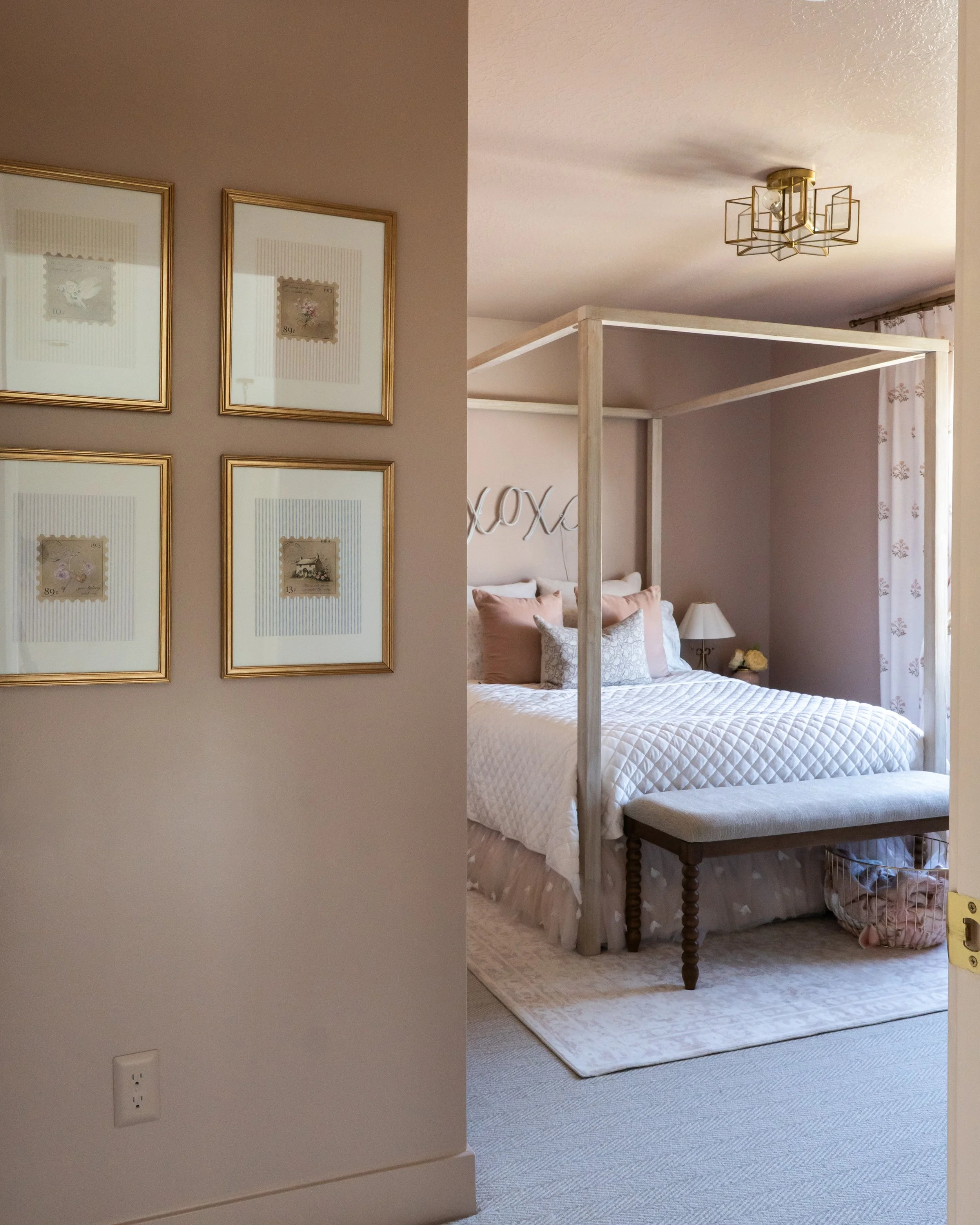 A view of a bedroom with a white bed, pink and white pillows, a side table with a lamp, and a canopy bed frame. The room has a beige wall, patterned curtains, and a ceiling light fixture.