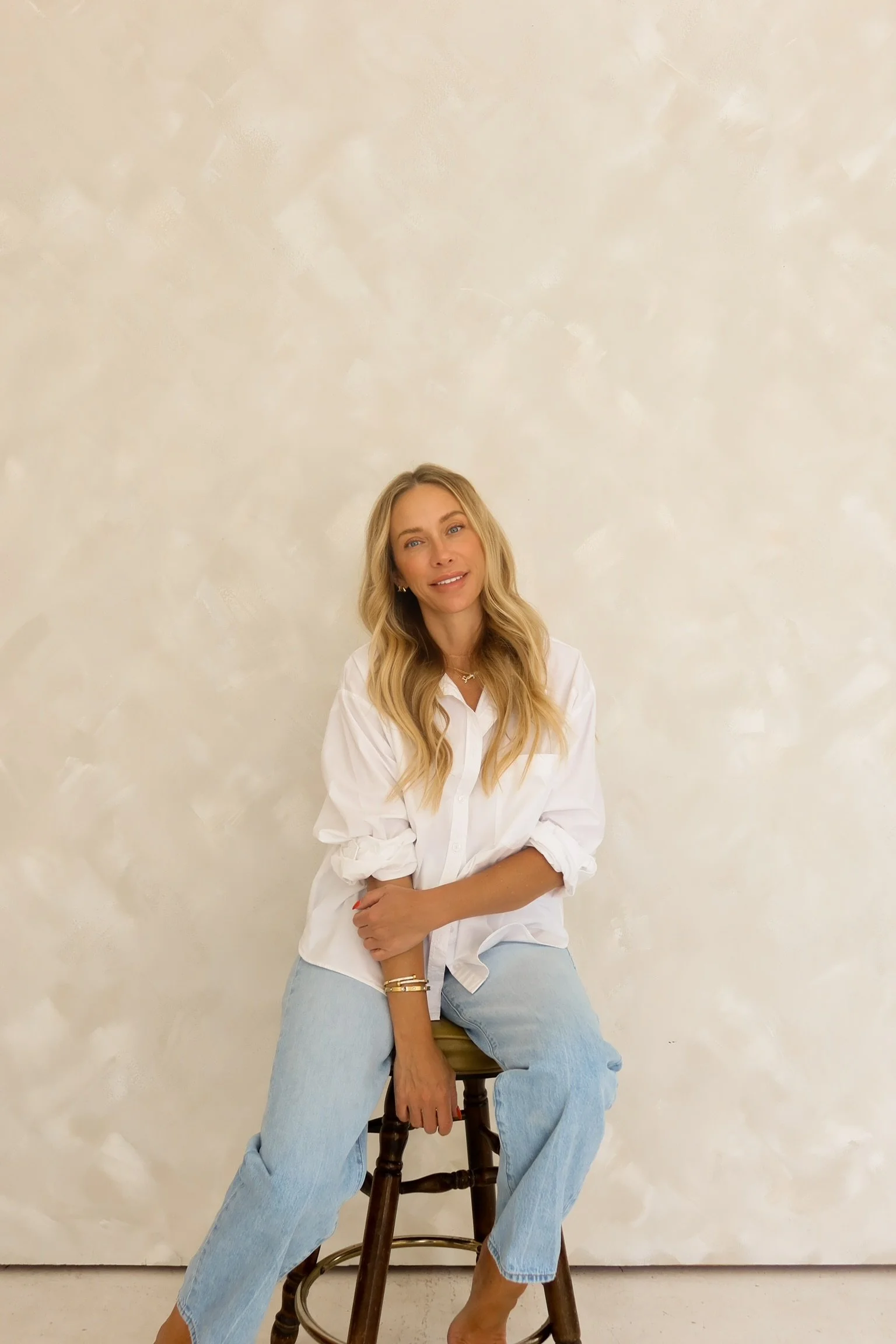 A young woman with long blonde hair, wearing a white shirt and light blue jeans, sitting on a wooden stool against a beige textured wall.