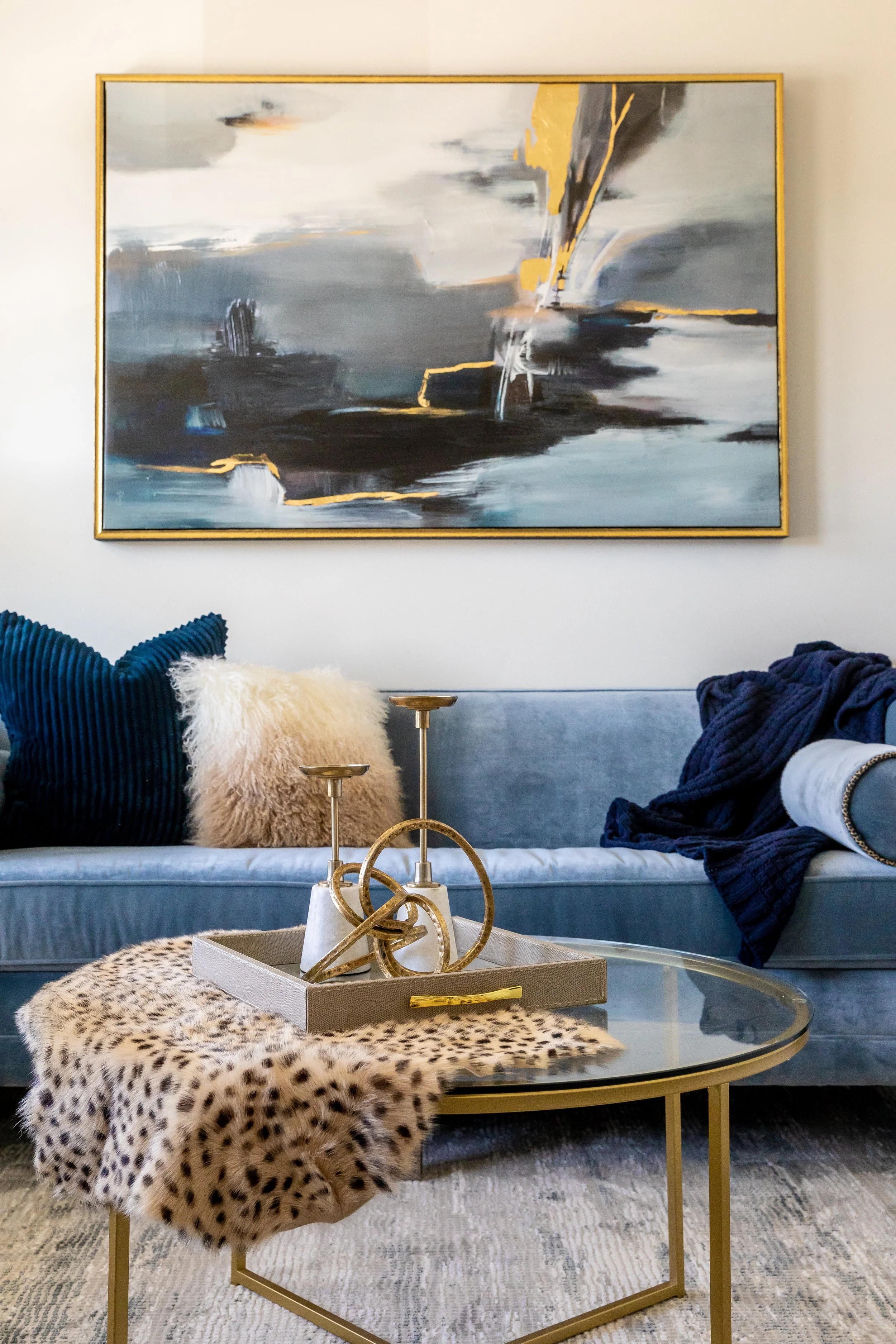 Living room wall art of an abstract painting with gray, white, black, and yellow tones, above a gray sofa with colorful textured pillows, a glass coffee table with a decorative gold sculpture, and leopard print and faux fur throws on the table and so