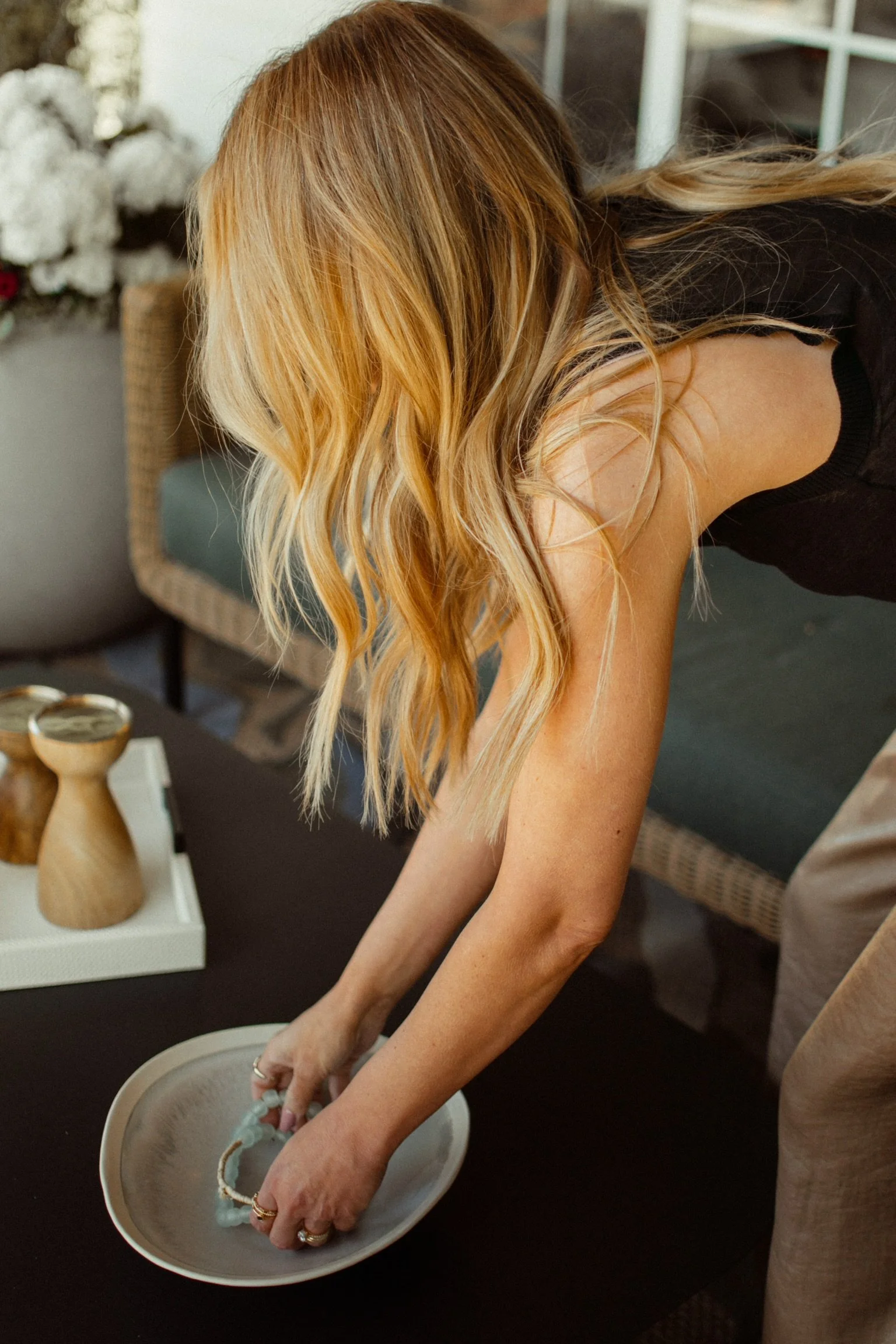 A woman with long, wavy blonde hair is placing her hands in a light-colored ceramic bowl on a dark table.