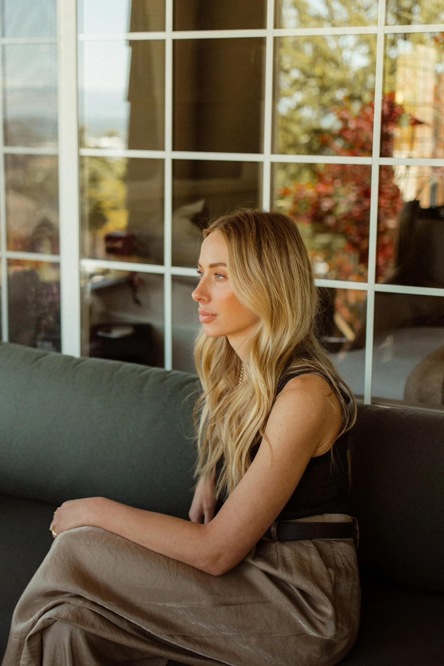 A woman with long blonde hair sitting on a dark green couch near a window with a grid pattern, with trees and a house visible outside