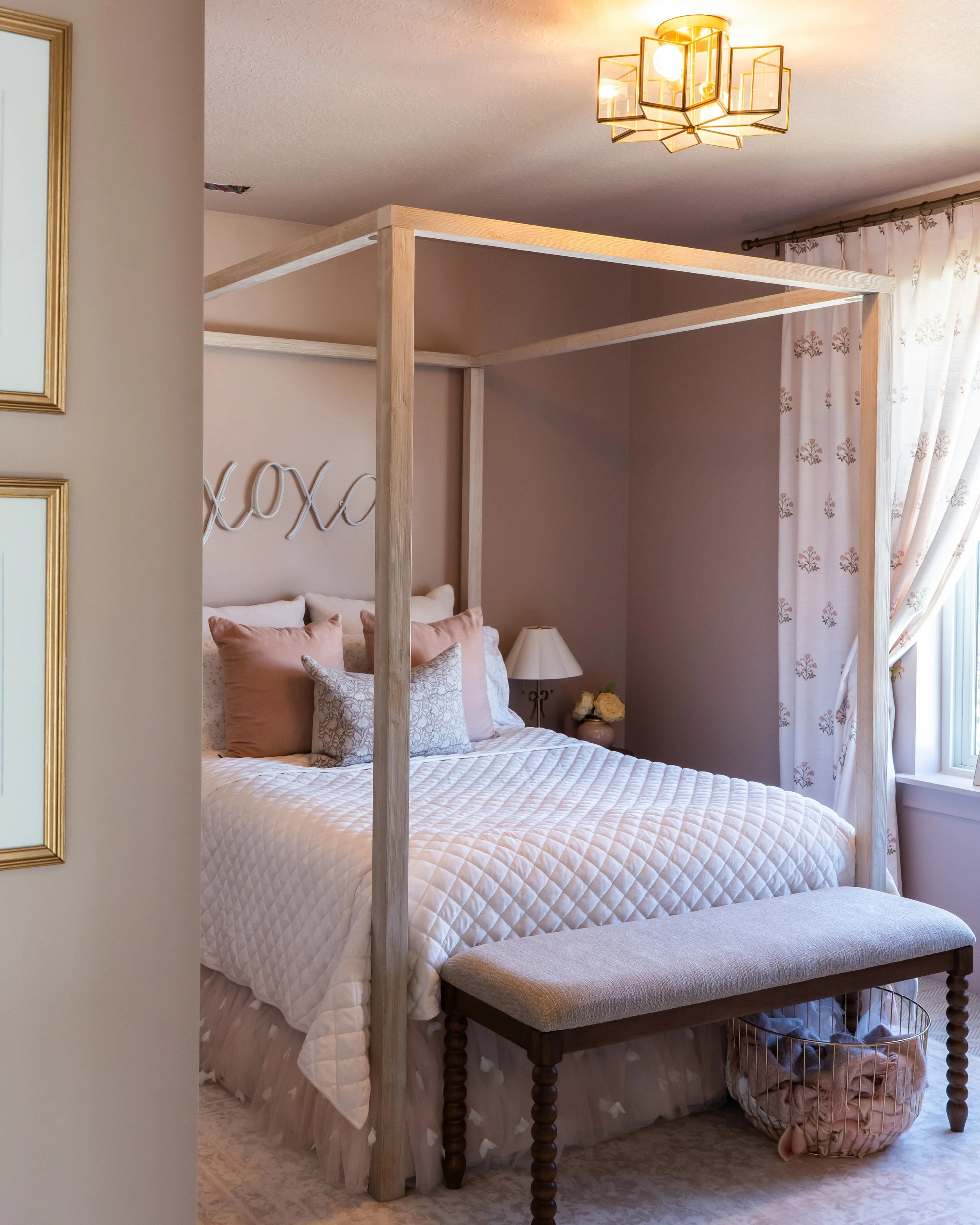 A cozy bedroom with a four-poster bed covered in a white quilt and blush pink pillows. Above the bed, there is a softly lit sign that reads 'xoxo.' To the right, there are white curtains with floral embroidery, and a window letting in natural light. 