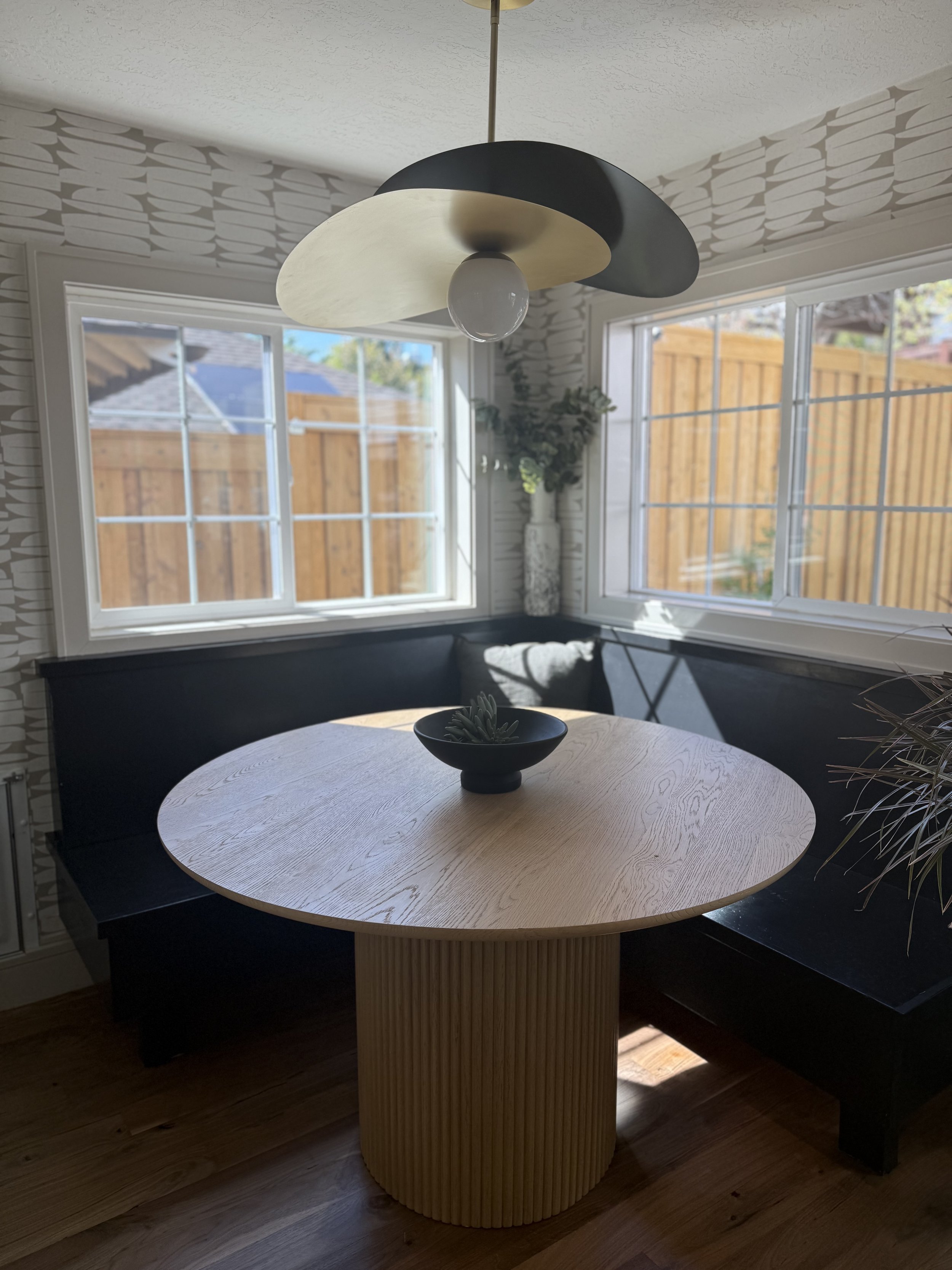 A round wooden dining table with a black curved bench on one side and a yellow vertical slatted support in the center. A black bowl with a succulent plant is on the table. The background has large windows showing a wooden fence outside, and a tall va