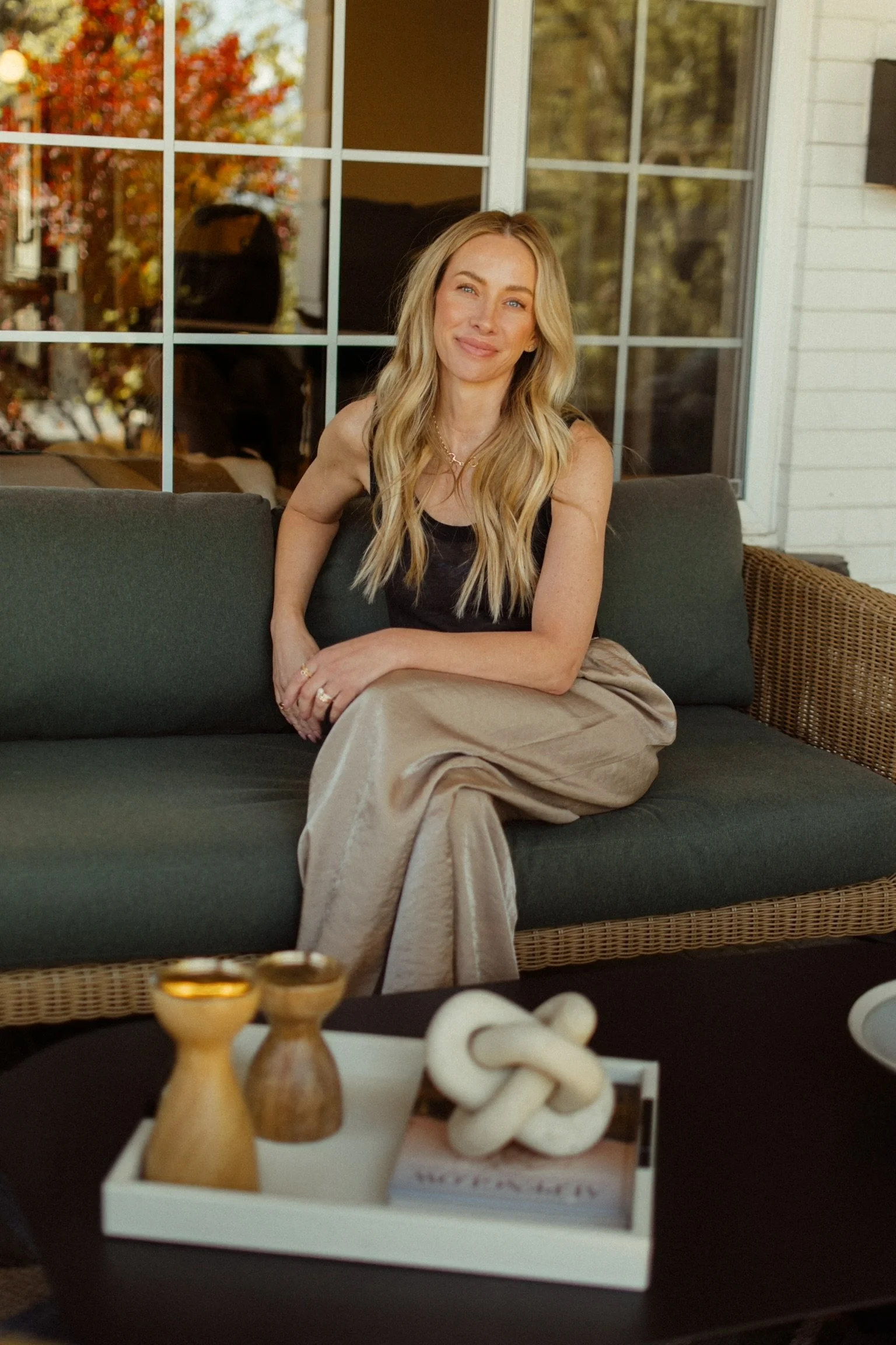 A woman sitting on a dark green outdoor sofa with a smile, blonde wavy hair, wearing a black top and beige pants, in a porch or patio area with trees outside.