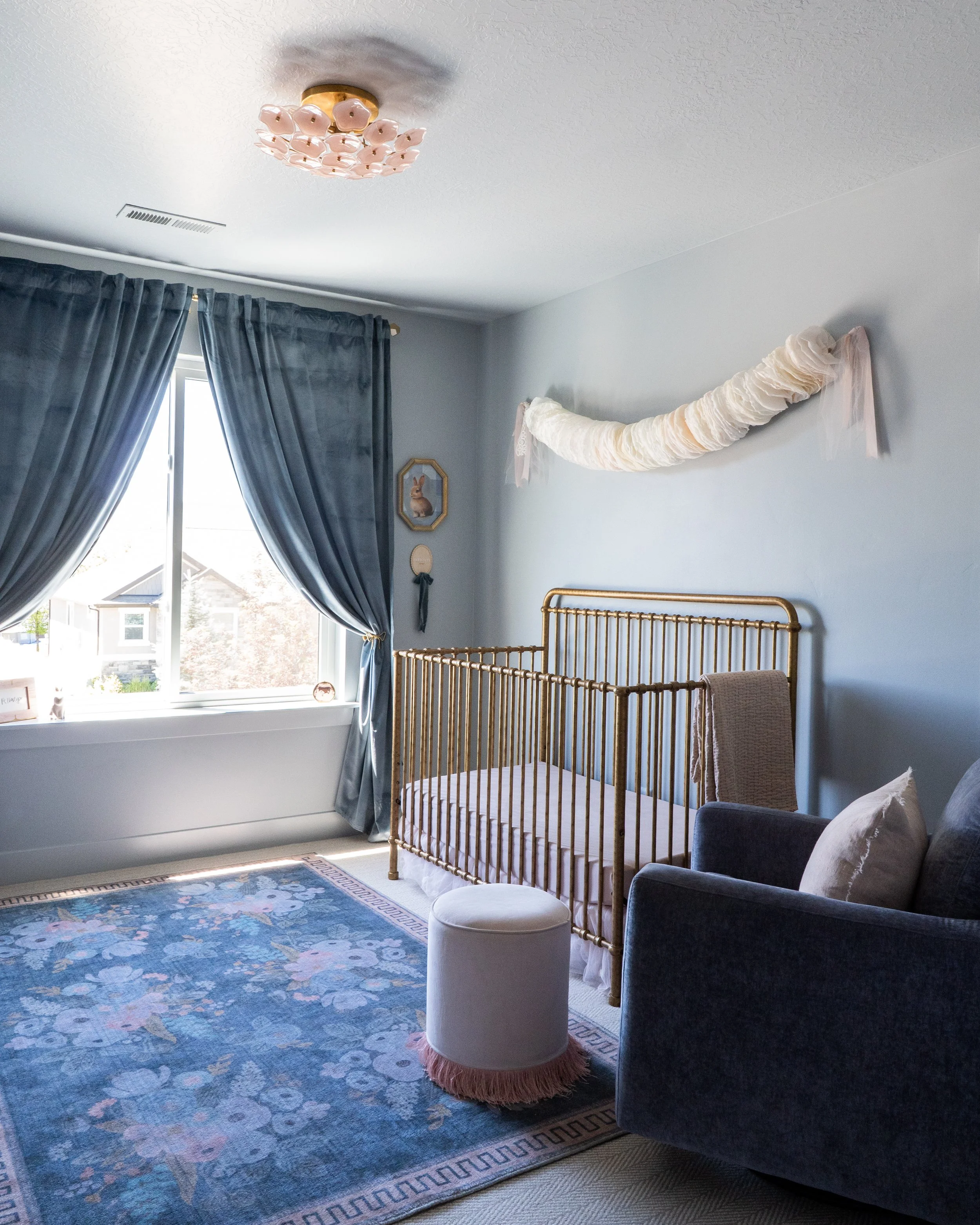 A nursery room with a blue floral rug, a dark upholstered armchair with a beige pillow, white and beige decorative fabric draped above a gold crib, and a window with blue curtains.