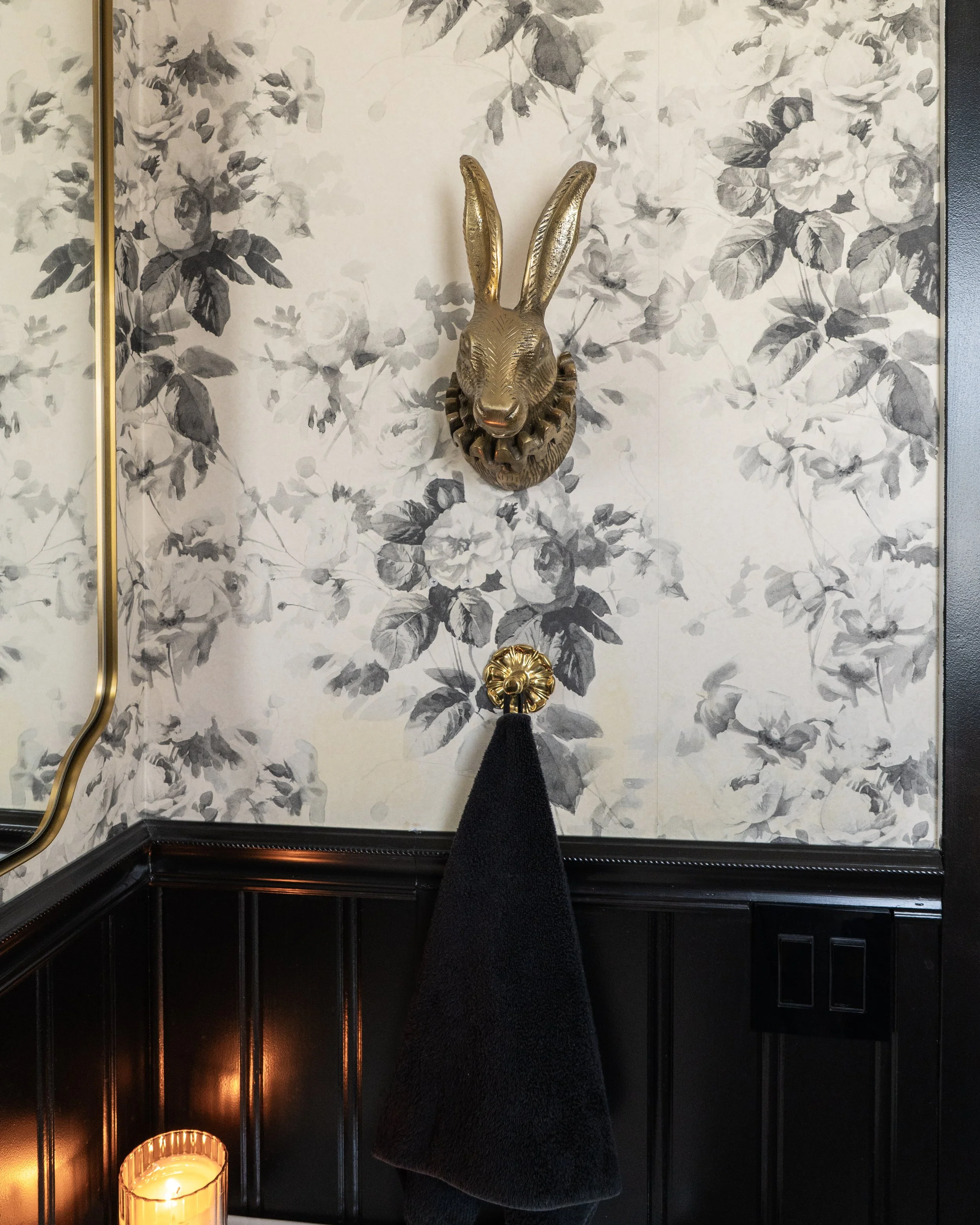 A gold decorative wall sculpture of a rabbit's head with long ears mounted on a wall with floral wallpaper. A black towel hangs below the sculpture, and there is a candle in a glass holder casting warm light in the bottom left corner.