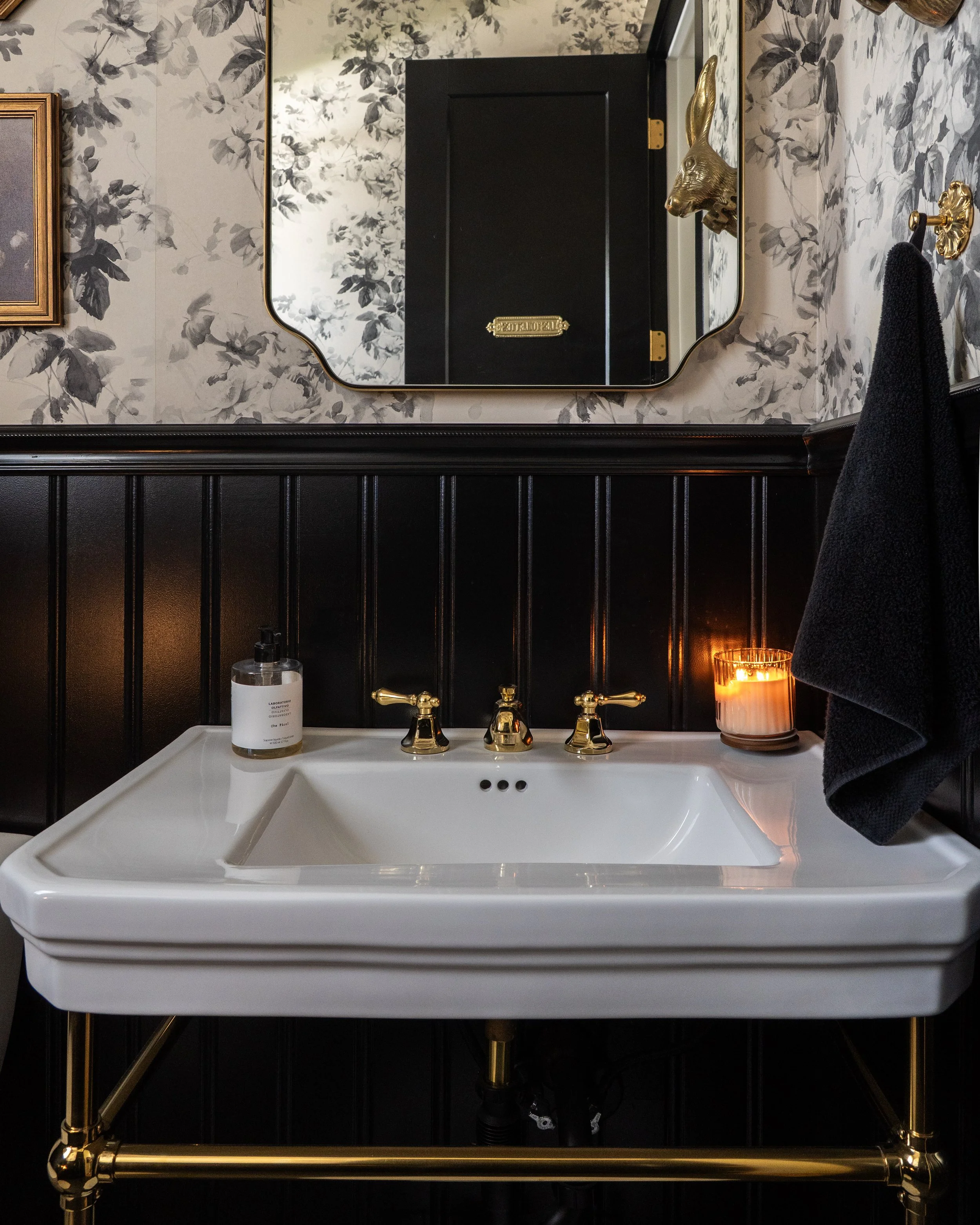 A bathroom vanity with a white sink, gold fixtures, and a black and gold framed mirror. The wall features black and white floral wallpaper, and a black towel hangs on a gold towel ring. On the sink, there is a bottle of liquid soap and a lit candle.