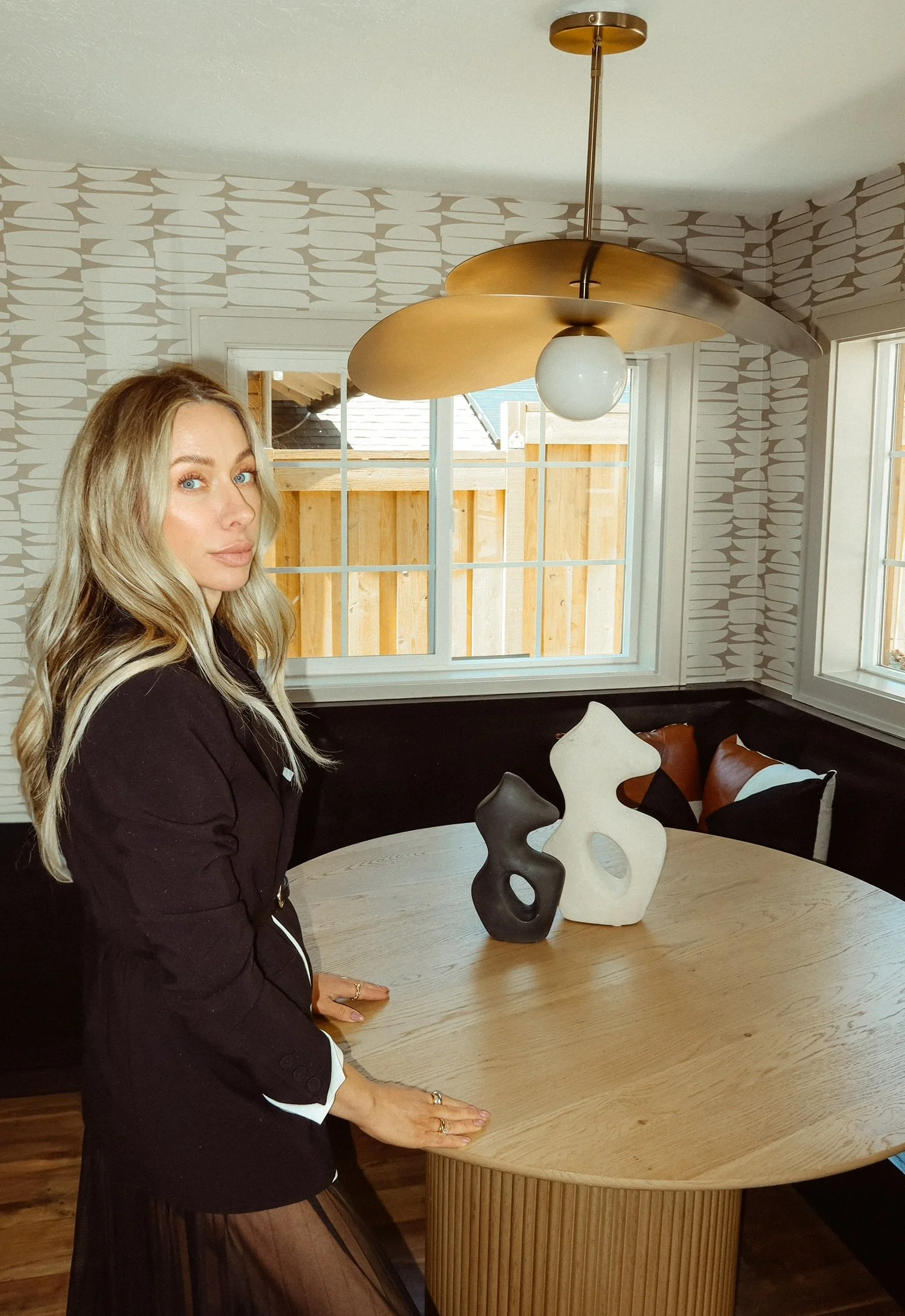 A woman with blonde hair and fair skin wearing a black jacket, sitting at a round wooden table with decorative vases, in a room with patterned walls and large windows.