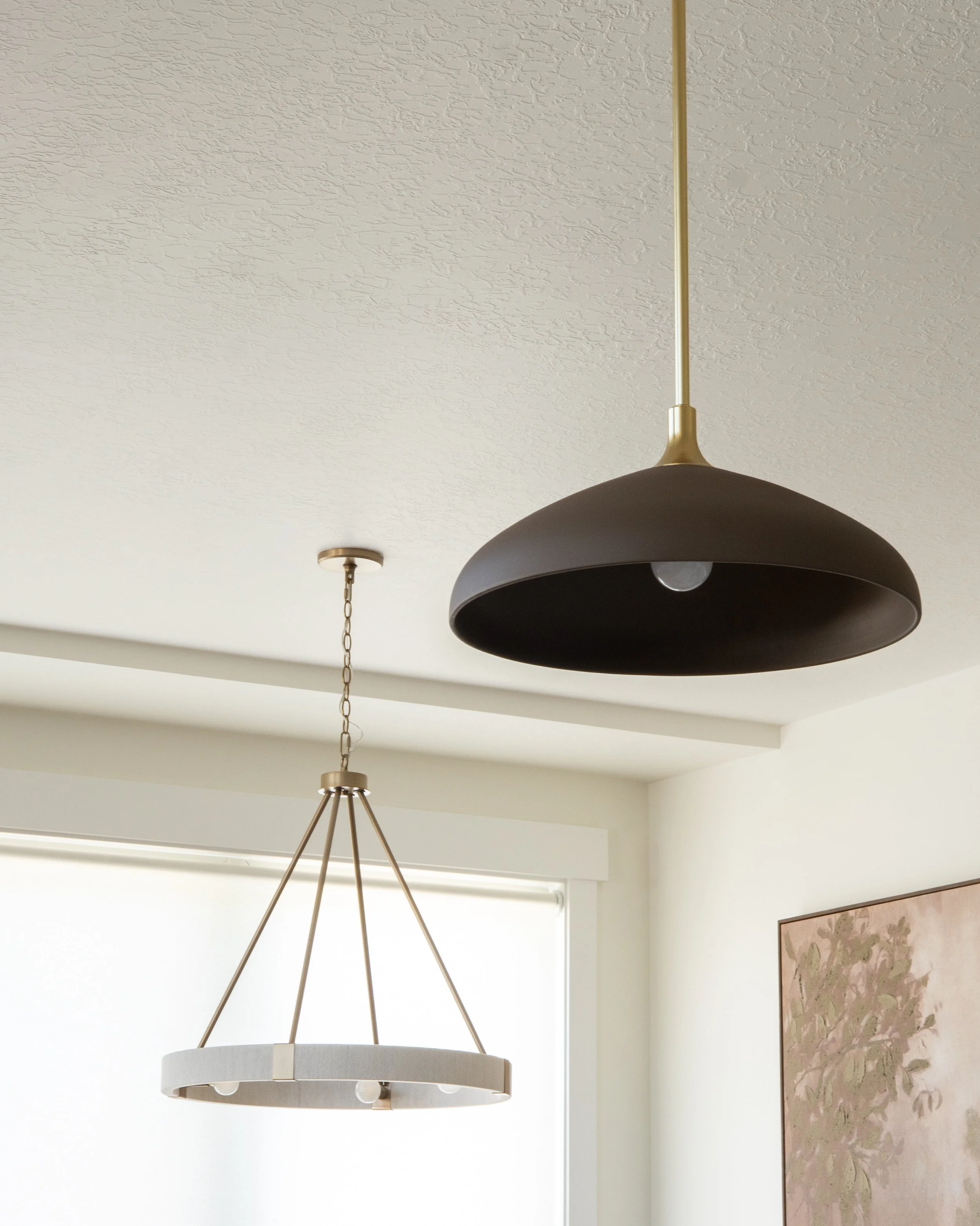 A modern black pendant lamp hanging from a white textured ceiling, with a small ceiling light fixture and a painting with floral patterns on the wall below.