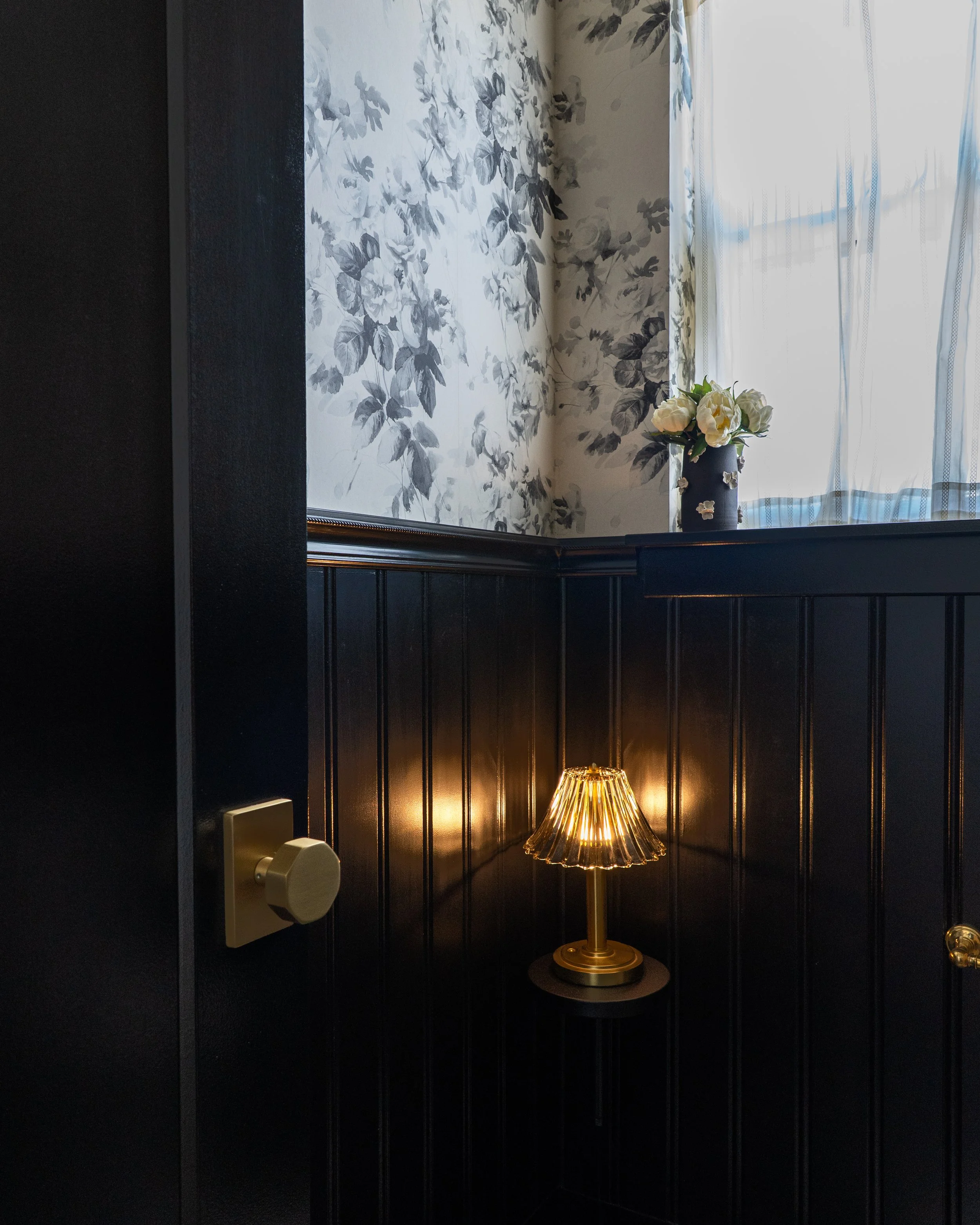 Inside a room with a dark wood-paneled wall and a floral wallpaper. There is a window with sheer white curtains, a dark-colored vase with white flowers, and a gold table lamp with a pleated shade on a round table.