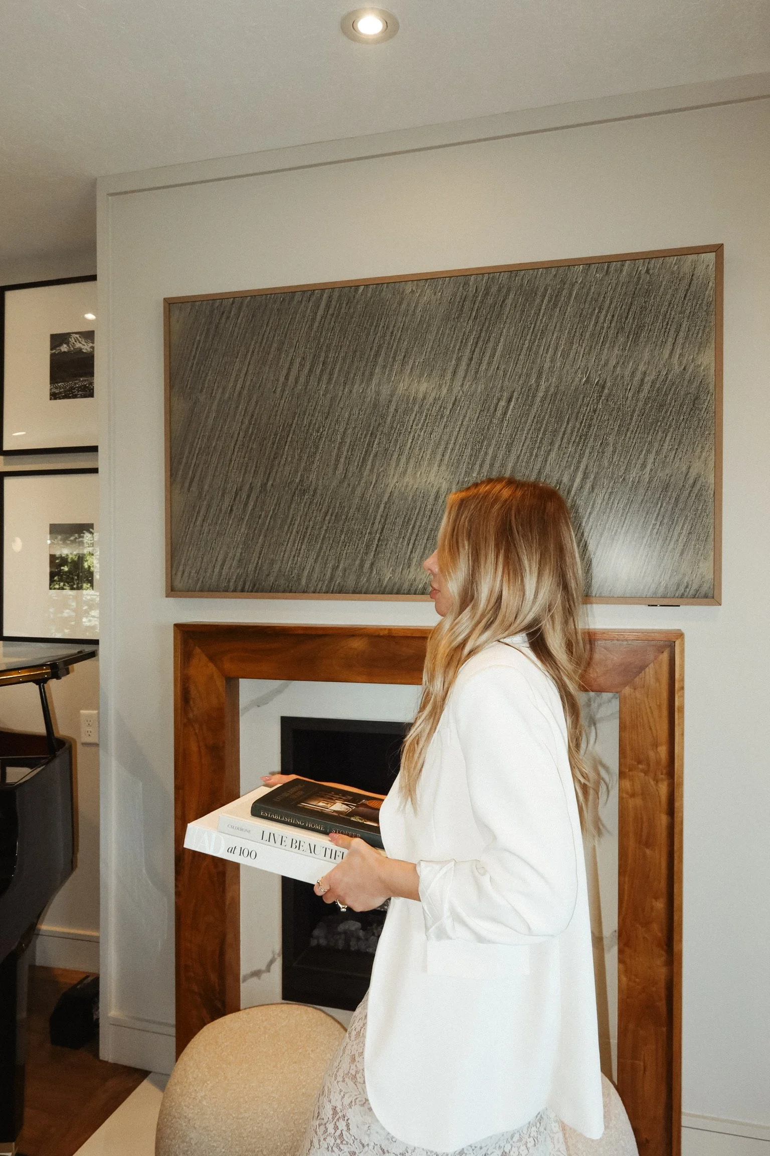 A woman with long blond hair wearing a white blazer and lace dress, holding a tray with magazines, standing in a living room with a fireplace and framed artwork on the wall.