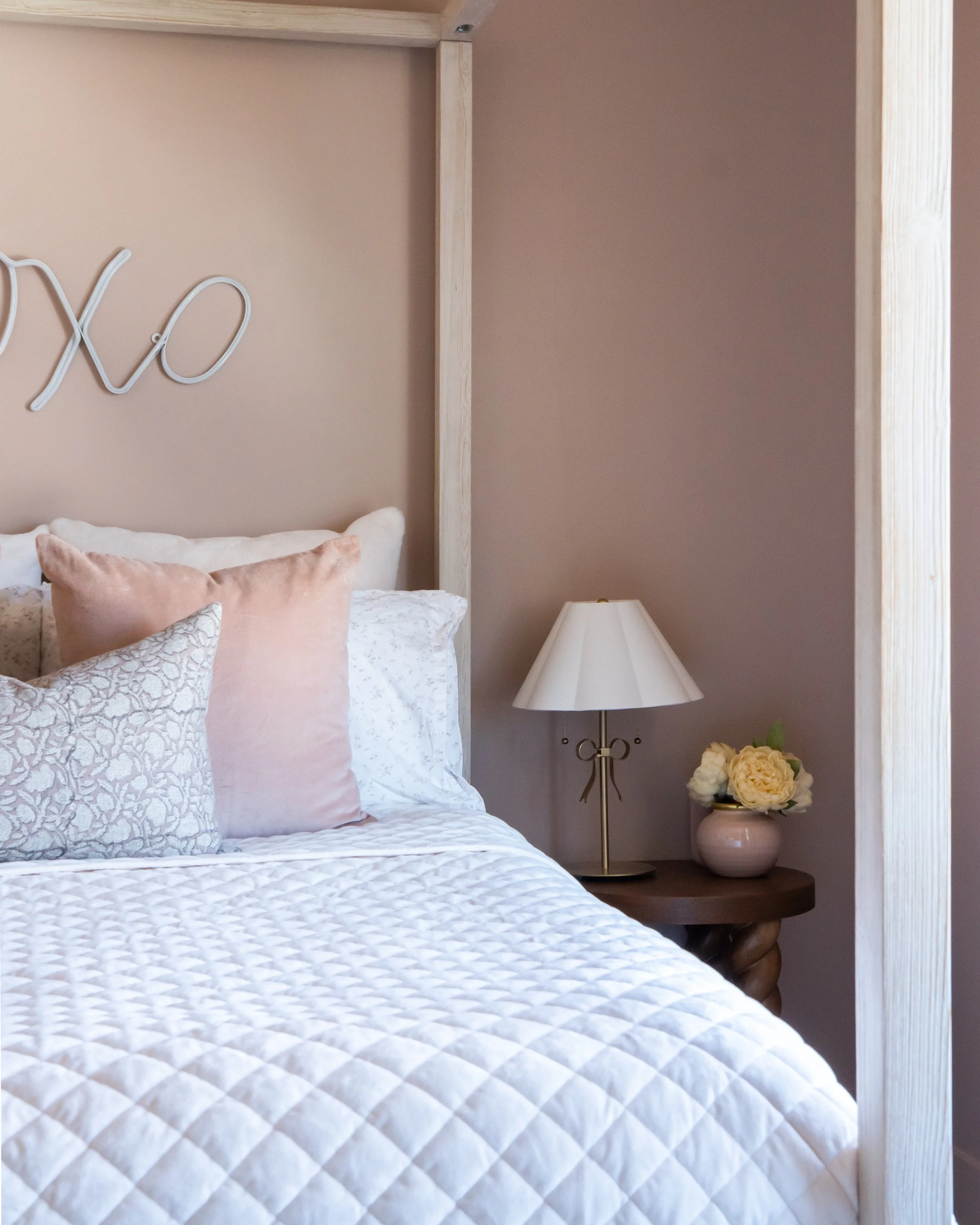 Close-up of a bed with white quilted bedding and multiple pillows in soft pink and patterned fabrics. Next to the bed is a wooden nightstand with a lamp and a pink vase with flowers. The wall behind is painted in a warm neutral color, and a wooden frame is partially visible.