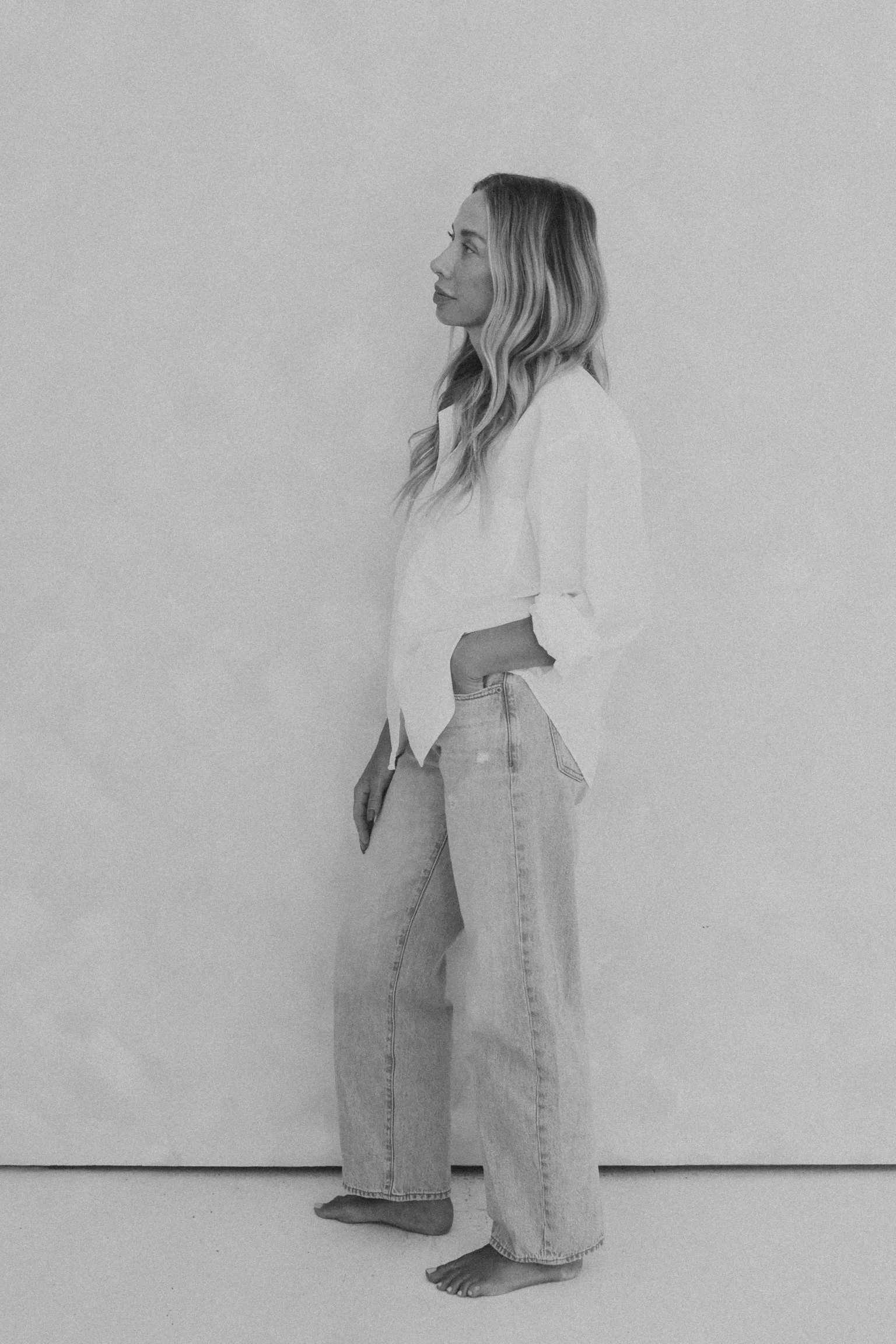 A woman standing barefoot on a plain surface, wearing a long, light-colored jeans and a loose shirt, with her hand in her pocket, and her hair styled with loose waves, captured in a side profile in a black-and-white photograph.