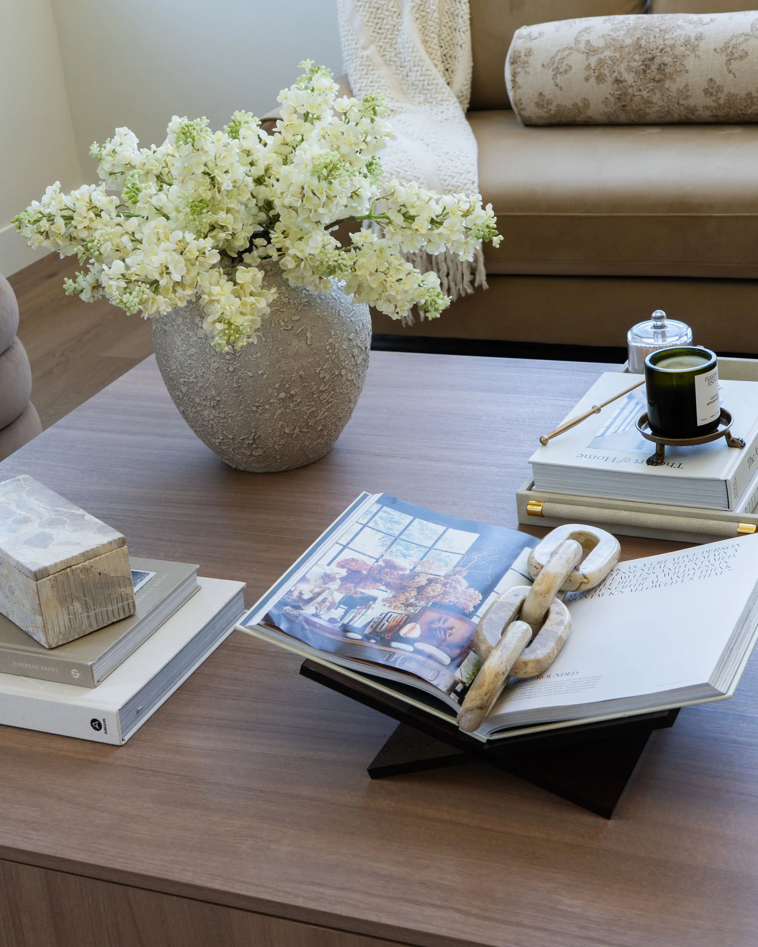 A wooden table with a large white vase filled with white flowers, alongside stacked books, a magazine, a wooden sculpture, a candle, and a small container with a lid. In the background, there is a beige sofa with patterned pillows and a throw blanket