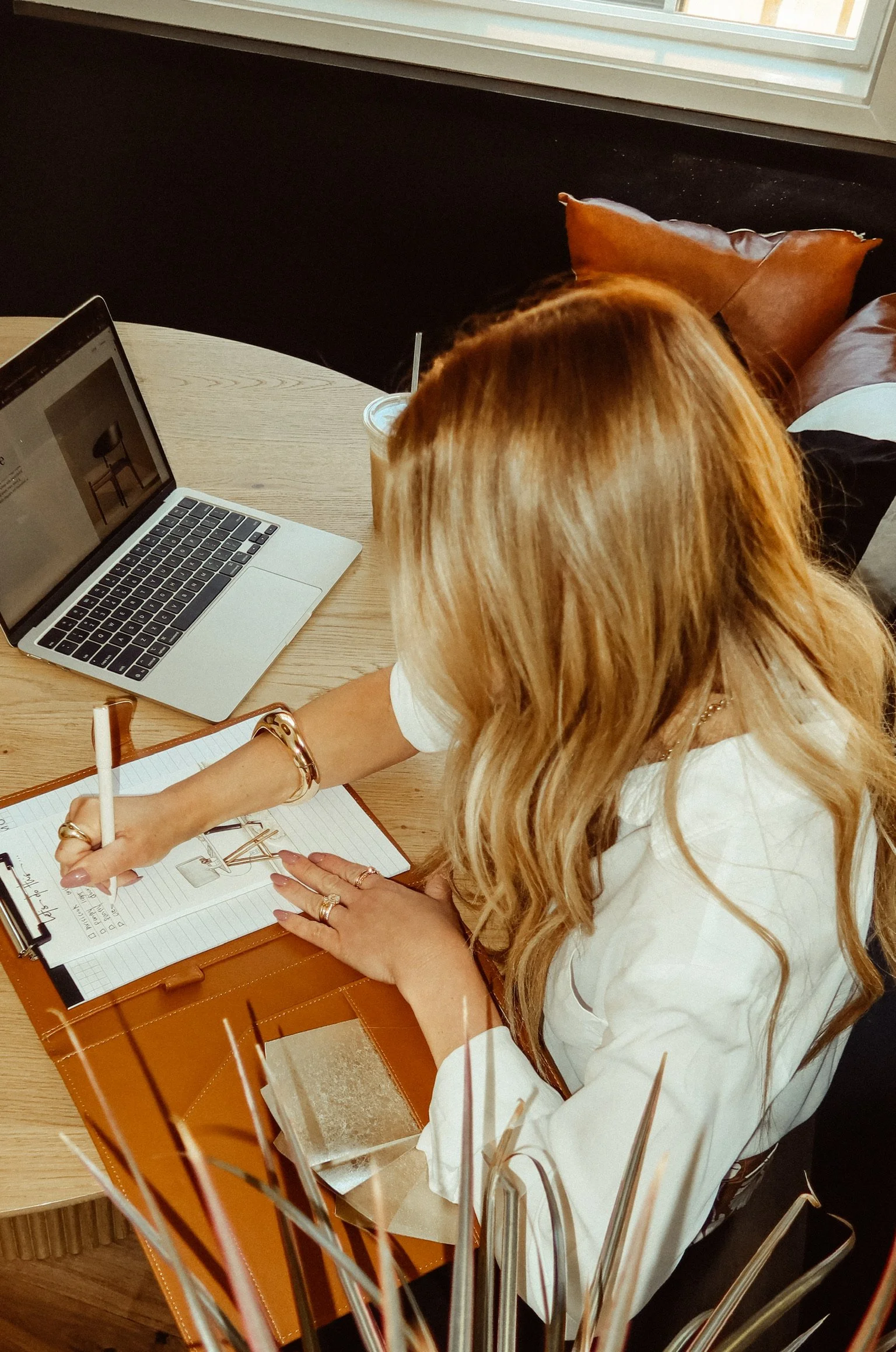 A woman with long, wavy blonde hair is sitting at a round wooden table, writing in a notebook with a pen. She is wearing a white blouse and multiple rings, and her left arm has a gold bracelet. On the table, there is an open leather planner, a MacBook laptop displaying a picture of a chair, and a drink with a straw. Behind her, there are brown pillows on a black cushioned bench near a window with natural light.