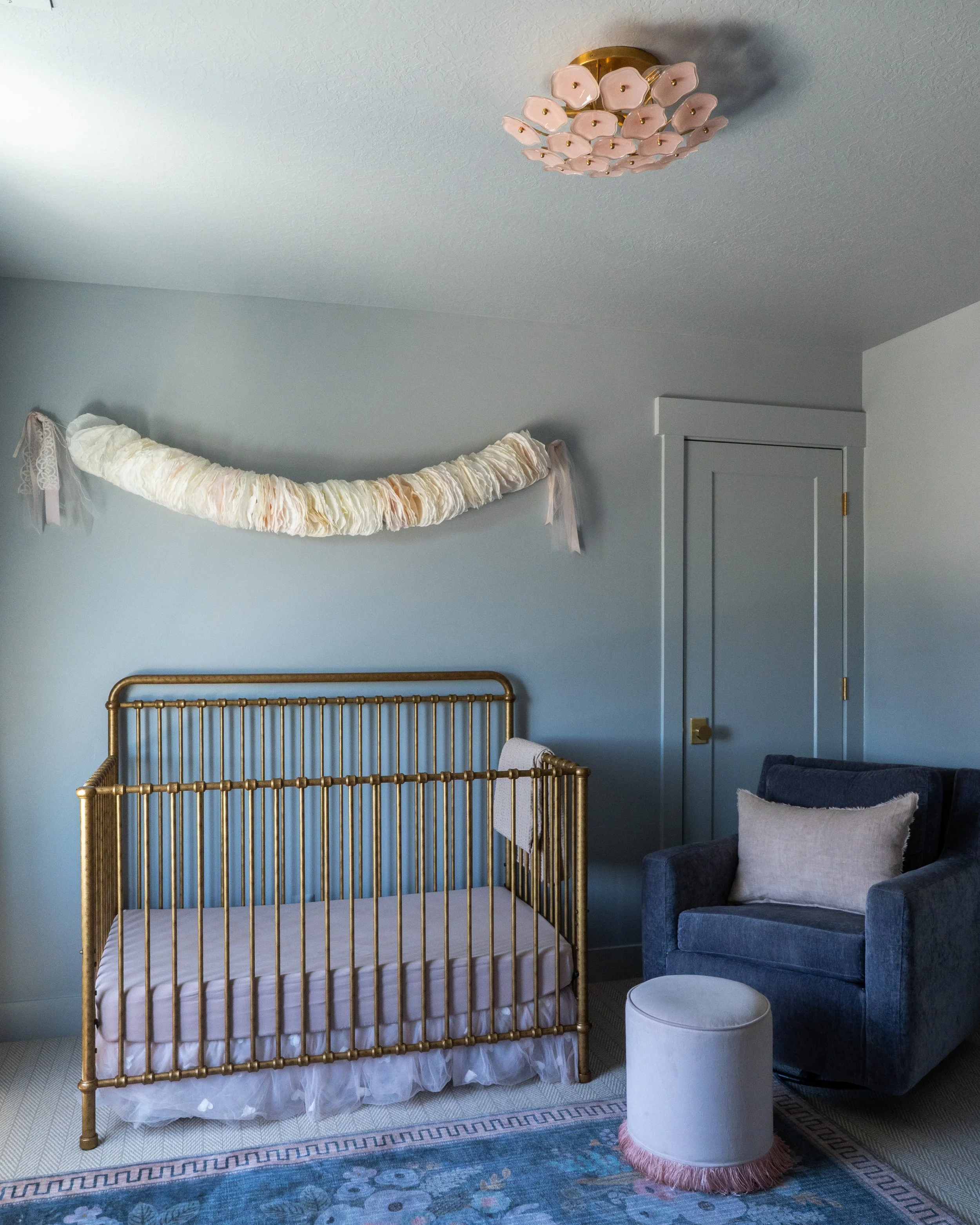 A bedroom with a brass bed frame, a small white round ottoman with pink fringe, a navy velvet armchair with a pillow, a gray door, and a pastel pink flower-shaped ceiling light. There is also a cream and pink fabric garland hanging on the wall.