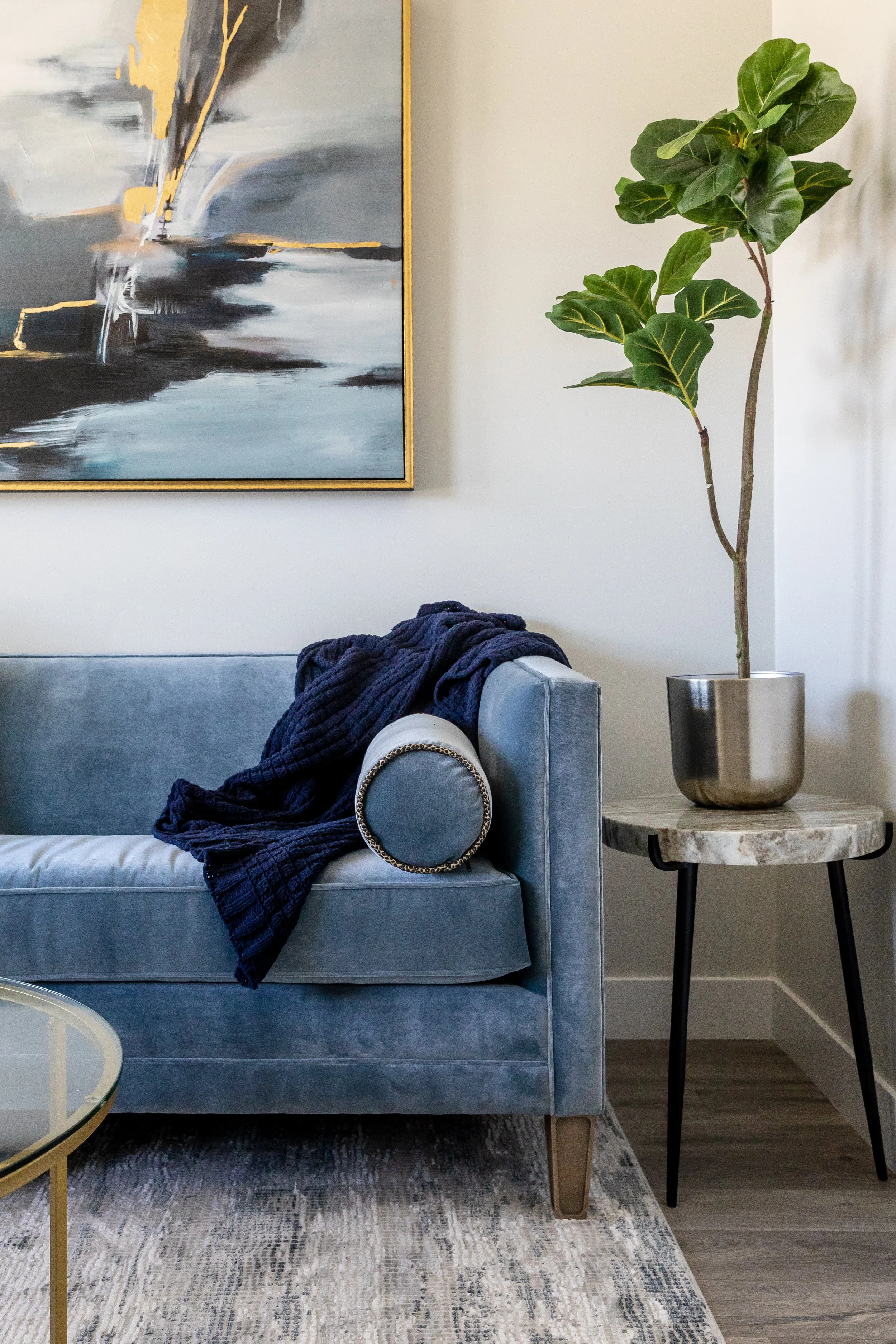 Interior of a living room featuring a light gray velvet sofa with a dark blanket draped over one arm, a round side table with a marble top and black legs, a potted fiddle leaf fig tree in a silver planter, a large abstract painting with black, white, gray, and gold tones on the wall, and a patterned area rug on the wooden floor.