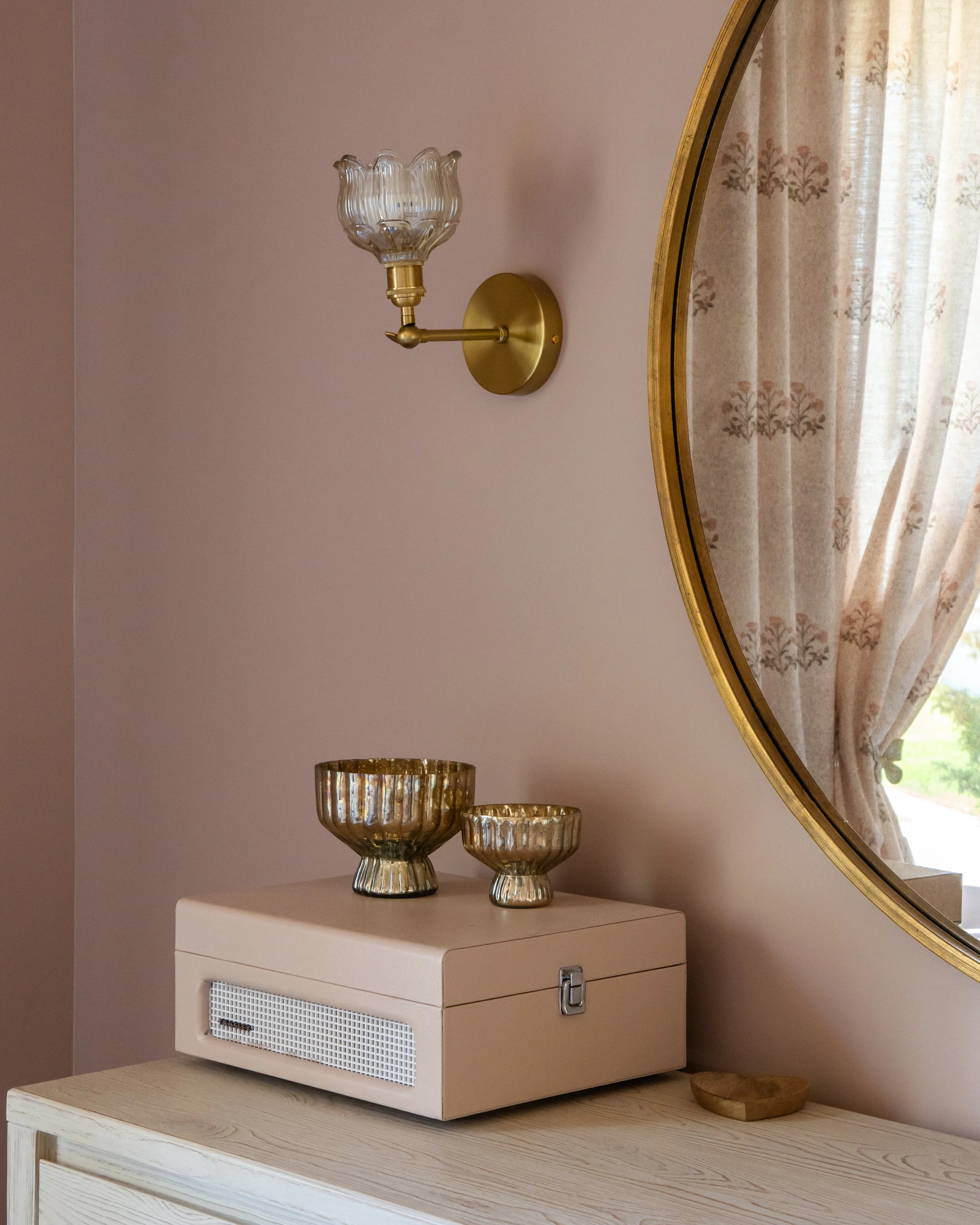 A beige vintage stereo or record player on a light wood dresser, with three gold glass bowls of varying sizes stacked on top, a round gold wall sconce, a gold-framed mirror, and a window with floral curtains reflected in the mirror.