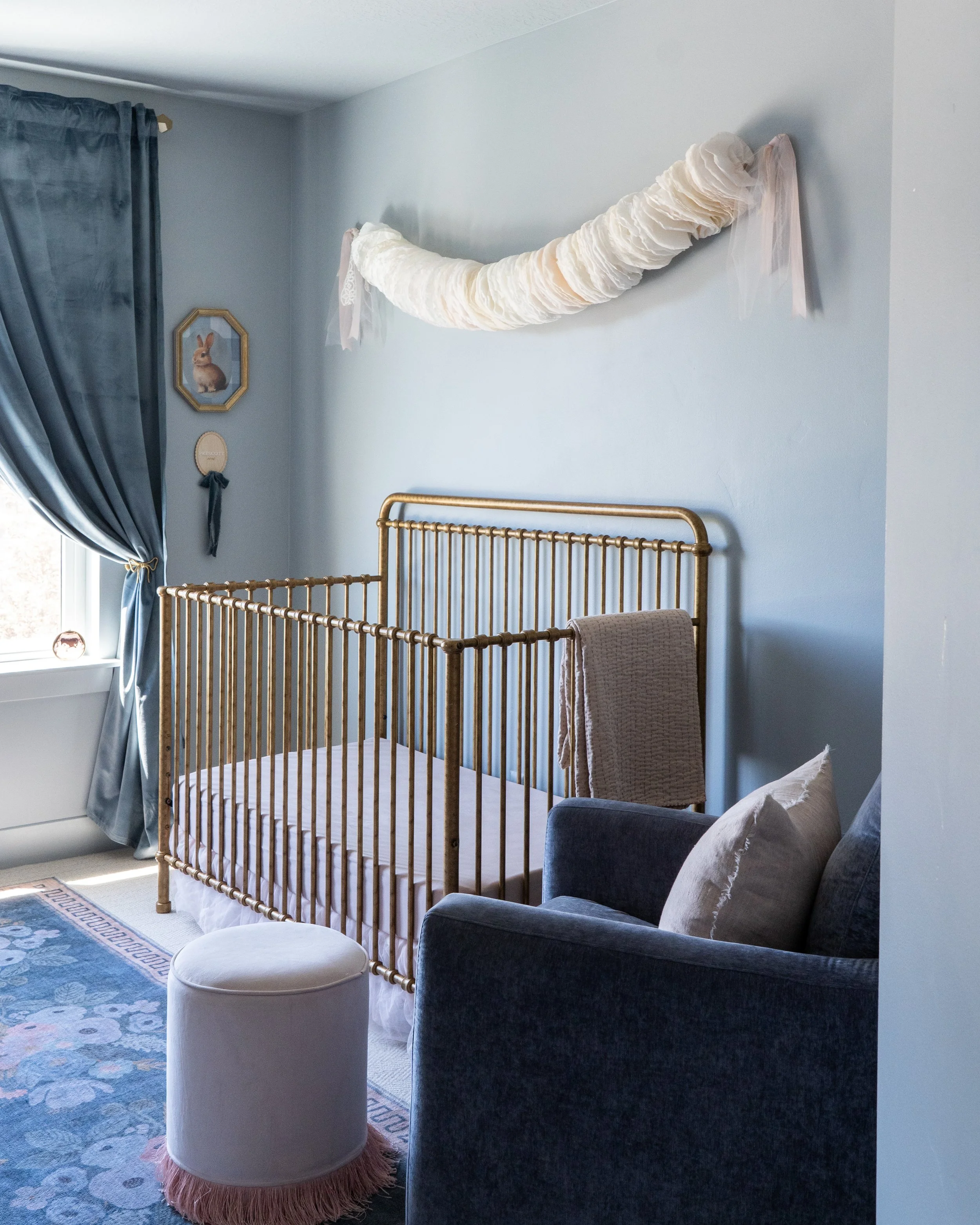 A bedroom with light blue walls, a brass bed frame, a navy blue armchair with pillows, a small white ottoman with a fringed edge, a blue floral area rug, and a window with navy curtains. There is also a wall decor featuring a rabbit