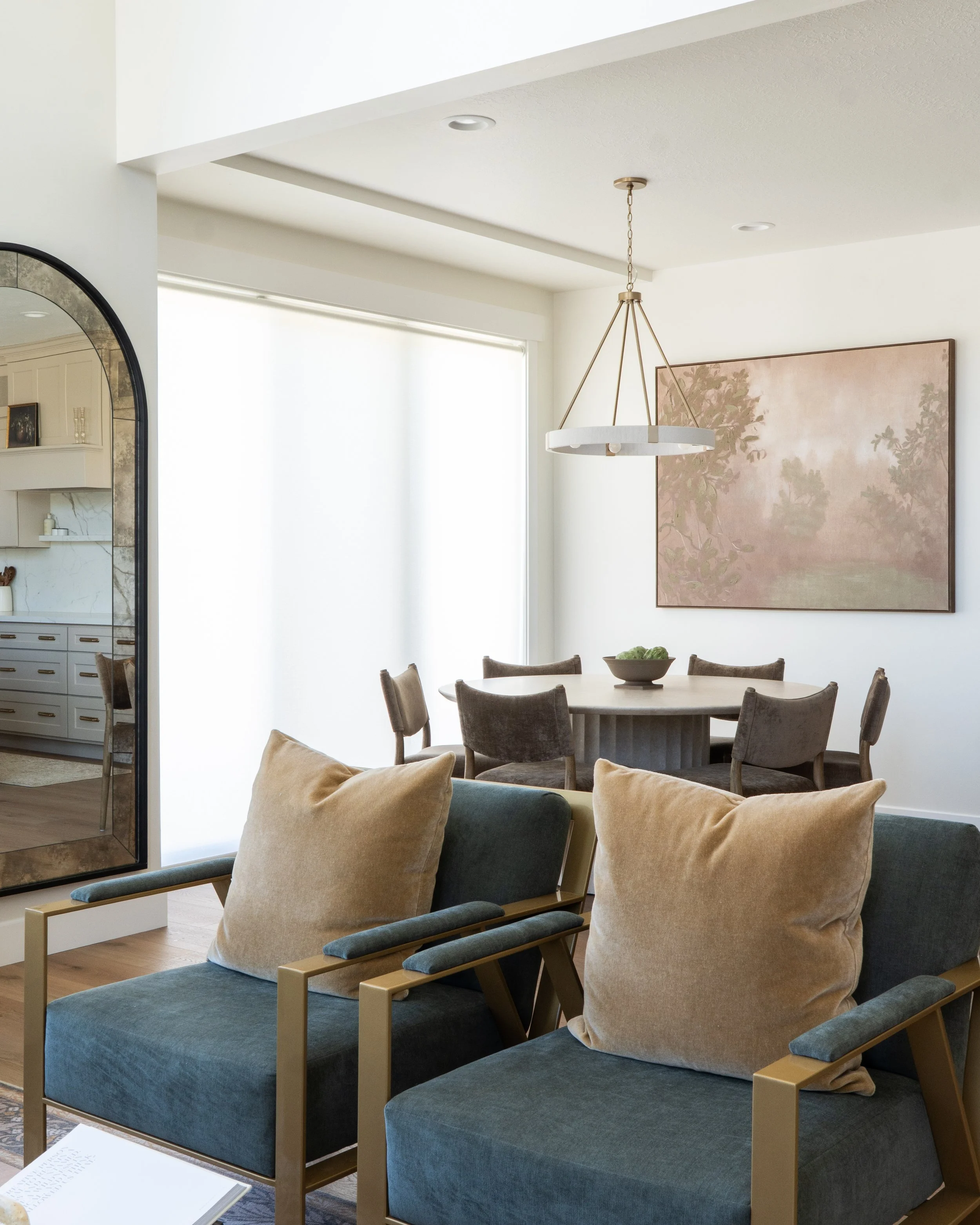 Living room with two blue chairs with beige pillows, a round table, and a large wall art of a pink landscape. A chandelier hangs from the ceiling, and a window with white blinds is visible.