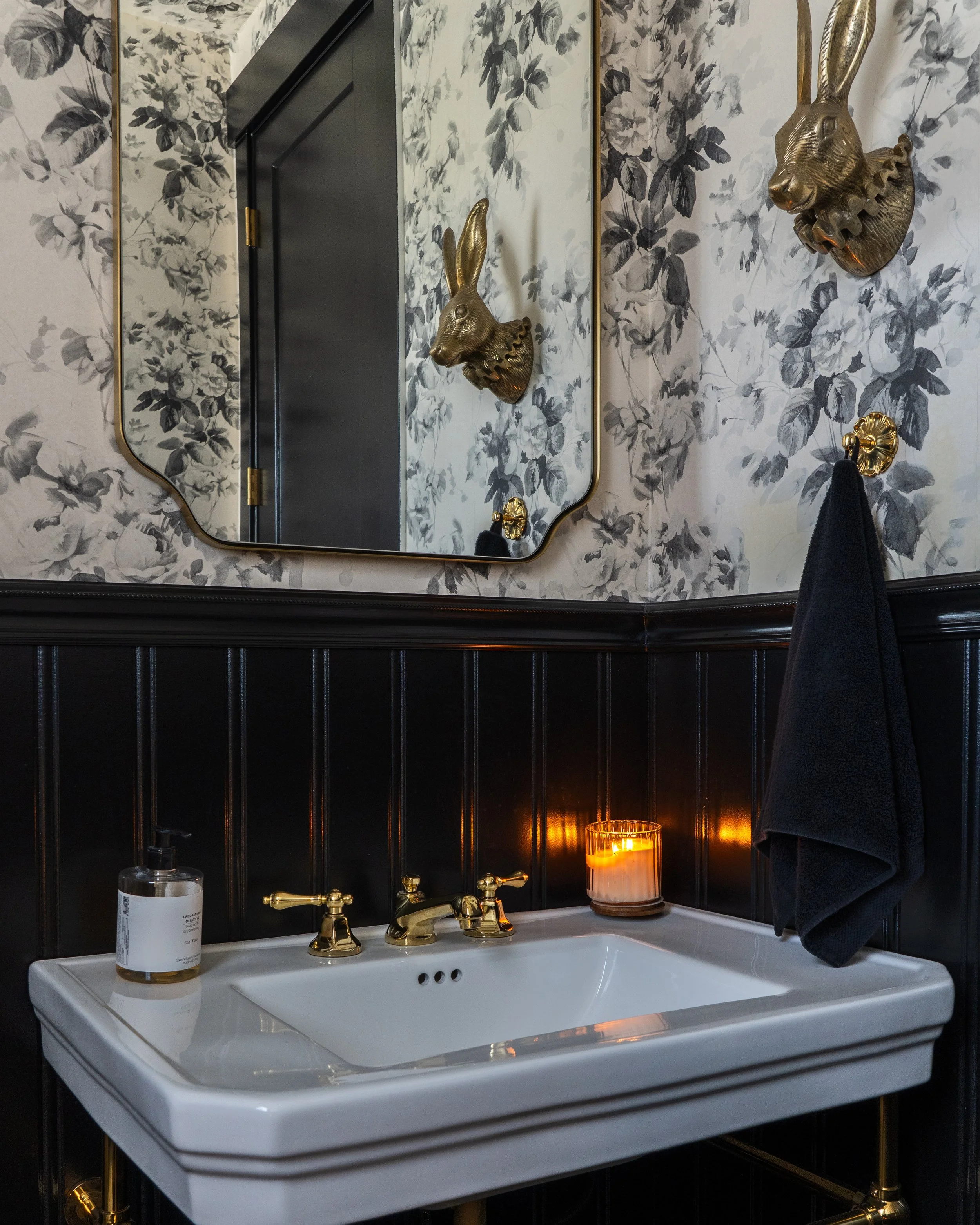 A vintage-style bathroom sink with gold fixtures, a large mirror, black and white floral wallpaper, a black towel, a lit candle, and a bottle of hand soap.
