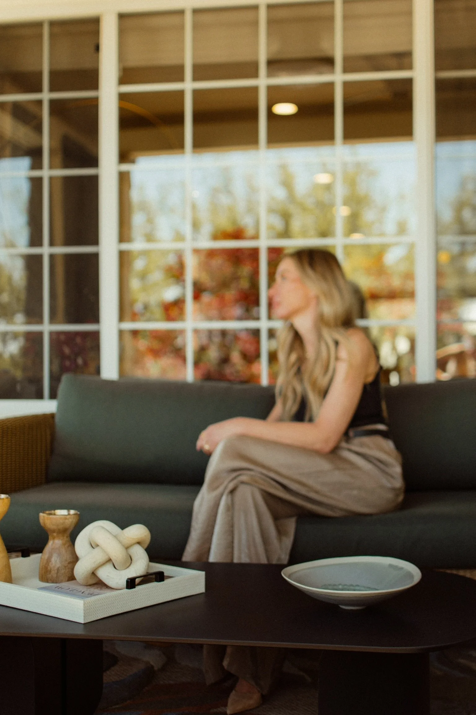 A woman with long hair sitting on a dark gray sofa inside a room with a large window, dressed in a black top and beige pants, with a warm, blurred autumn scene visible outside the window. In the foreground, a black coffee table holds a decorative whi