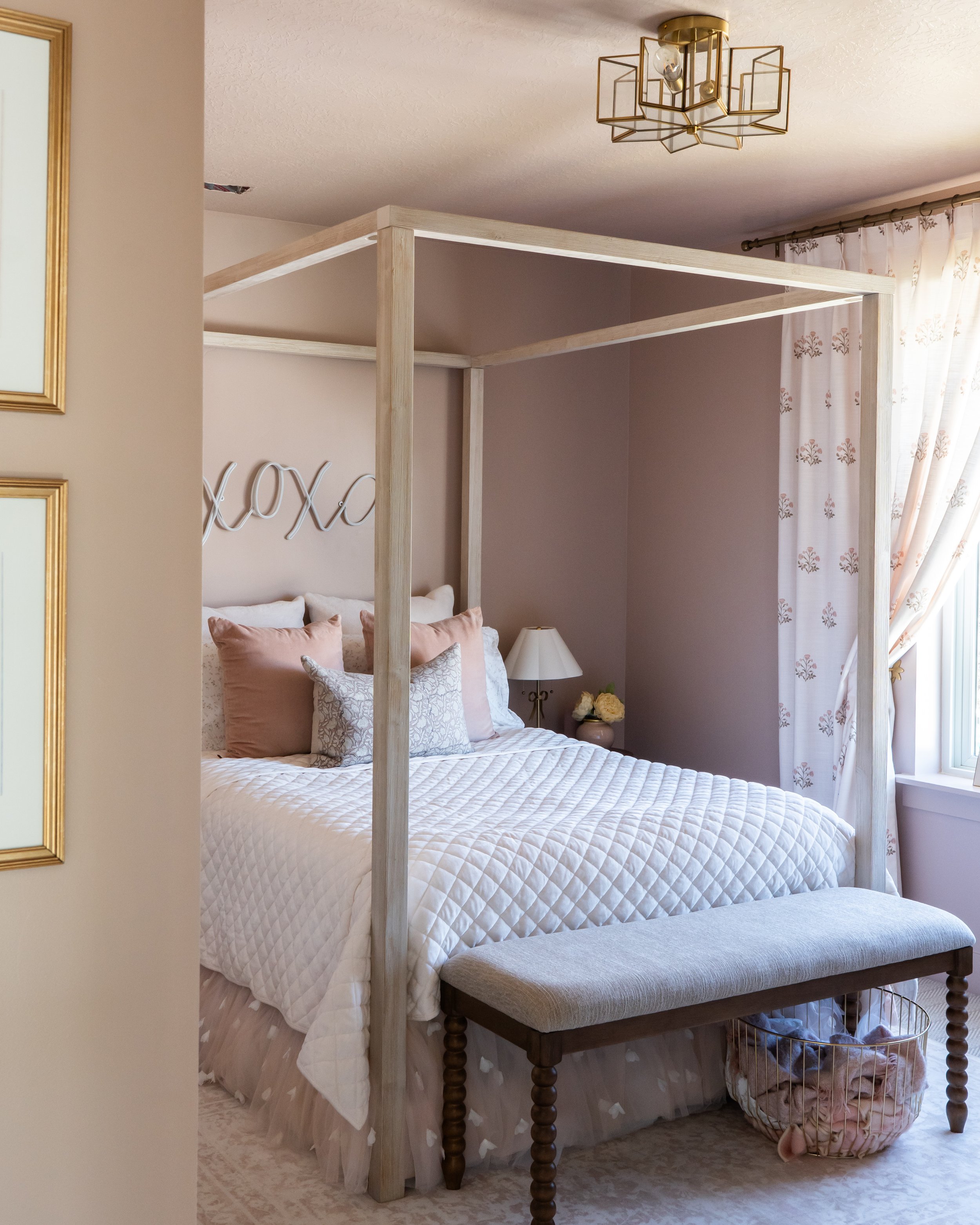 A cozy bedroom with a white bed covered in a textured quilt, decorated with soft pink and white pillows. A small table lamp is on the nightstand next to the bed. Above the bed, the word 'xoxo' is displayed on the wall. There is a wooden four-poster b