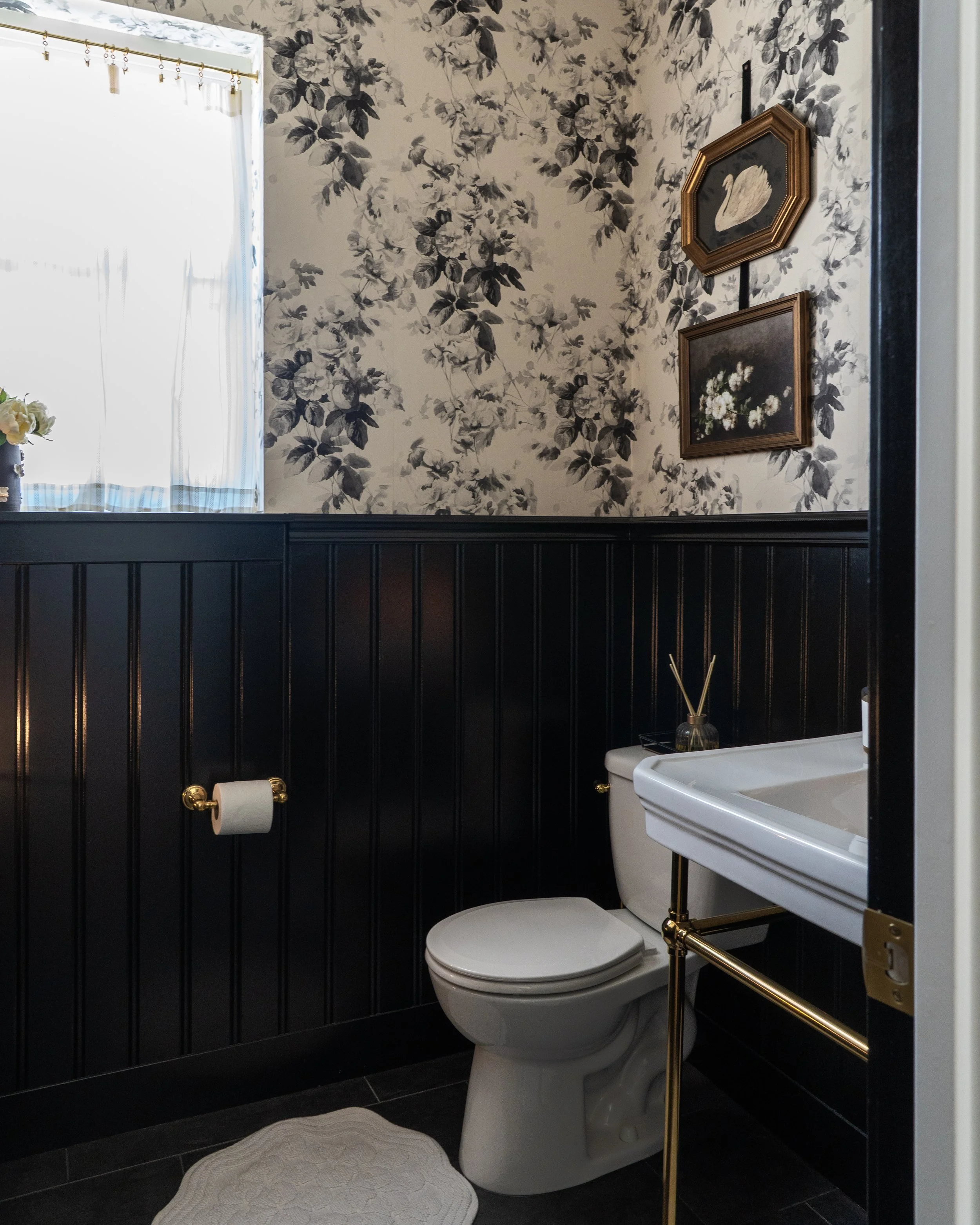 A compact bathroom with dark paneled lower wall and patterned floral wallpaper on the upper wall. The bathroom features a white toilet with a closed lid, a white sink with brass fixtures, and a small round rug on the dark tiled floor. A window with s
