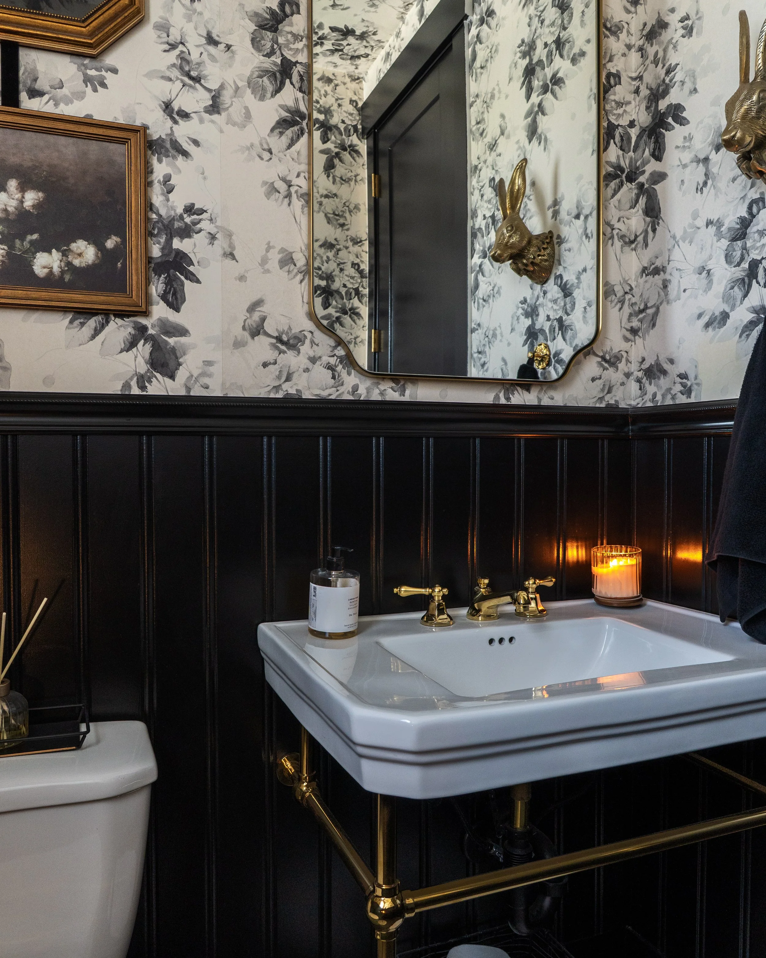 A bathroom with floral wallpaper, a black-framed mirror, and gold animal head wall sculptures. A white sink with gold fixtures, a candle, and a bottle of hand soap are visible below the mirror.