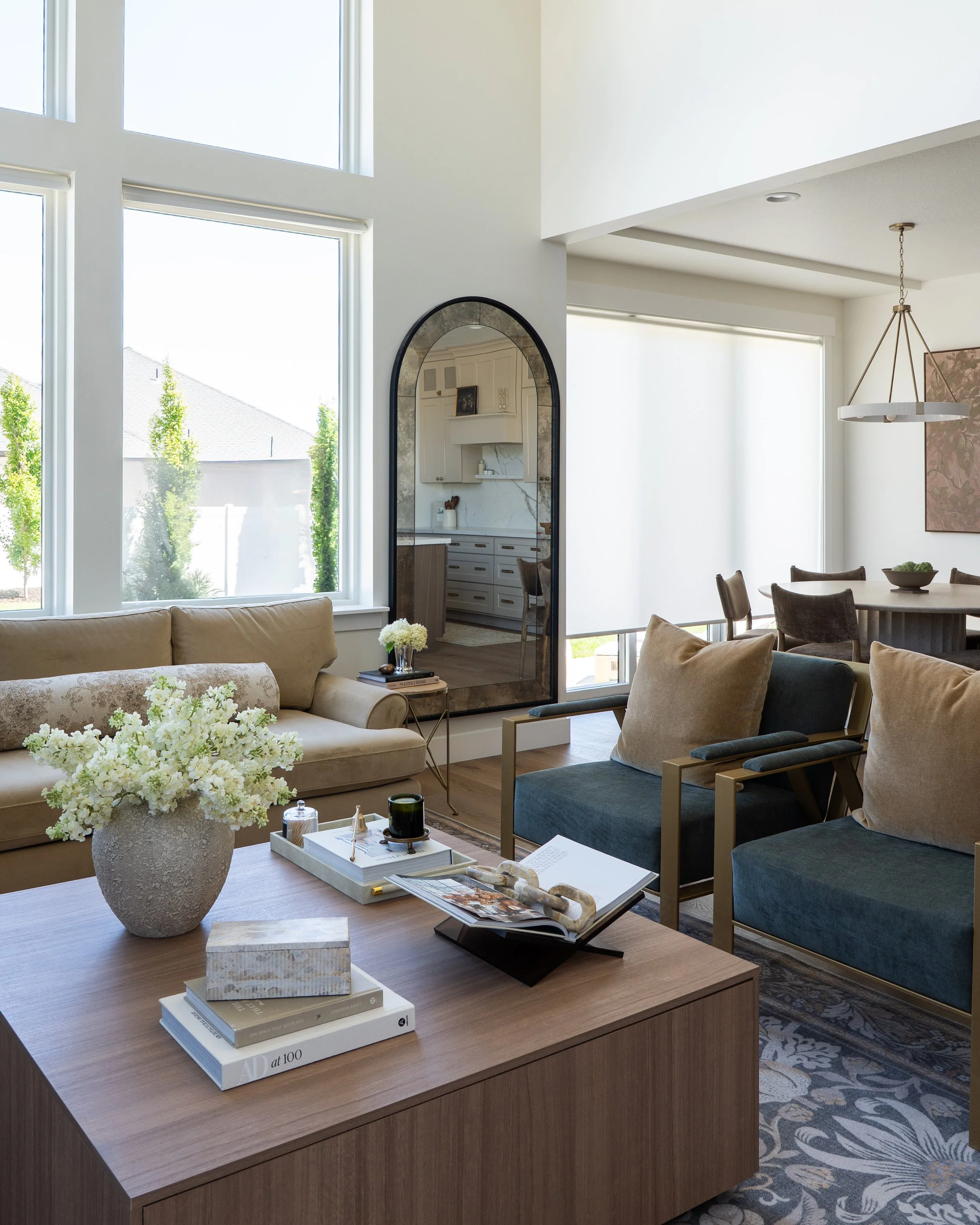 Modern living room with large windows, beige and blue armchairs, a wooden coffee table with books and decor, and a round floral arrangement in a textured pot.