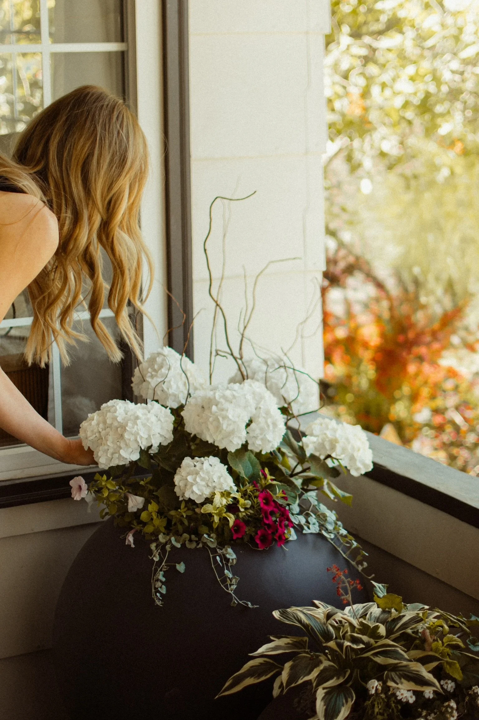 A woman with long, wavy blonde hair arranging white hydrangea flowers on a patio table next to a window, with fall-colored trees in the background.