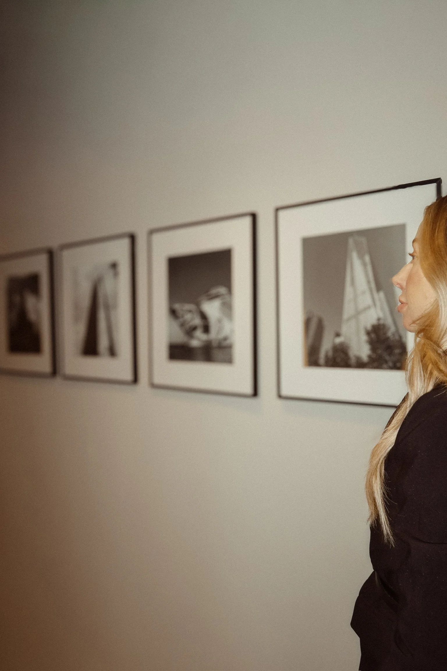 A woman with long blonde hair wearing a black outfit is looking at black and white photographs displayed on a gallery wall.