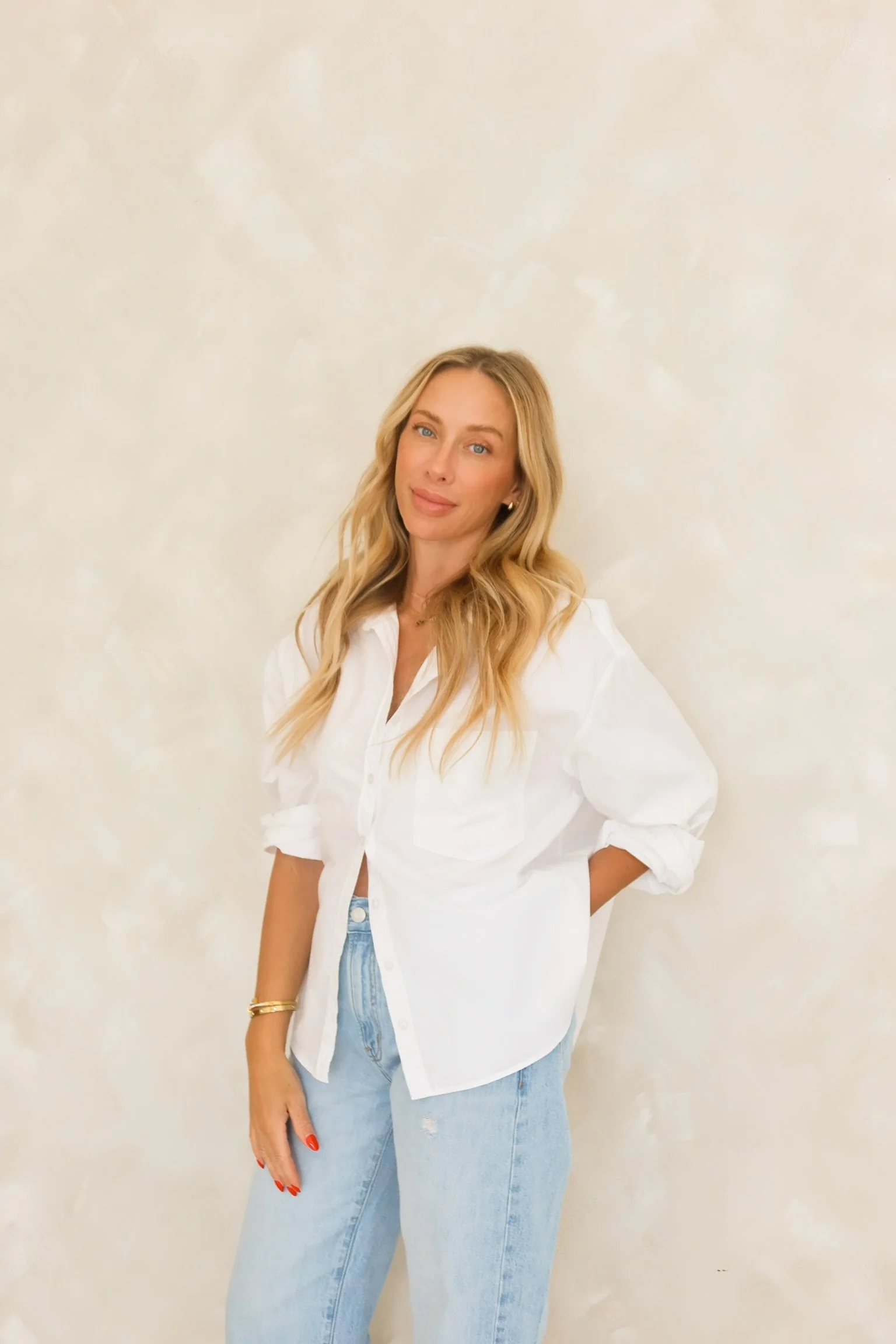 A woman with long blonde hair, wearing a white shirt, light-wash jeans, and gold jewelry, stands against a cream-colored, textured background.