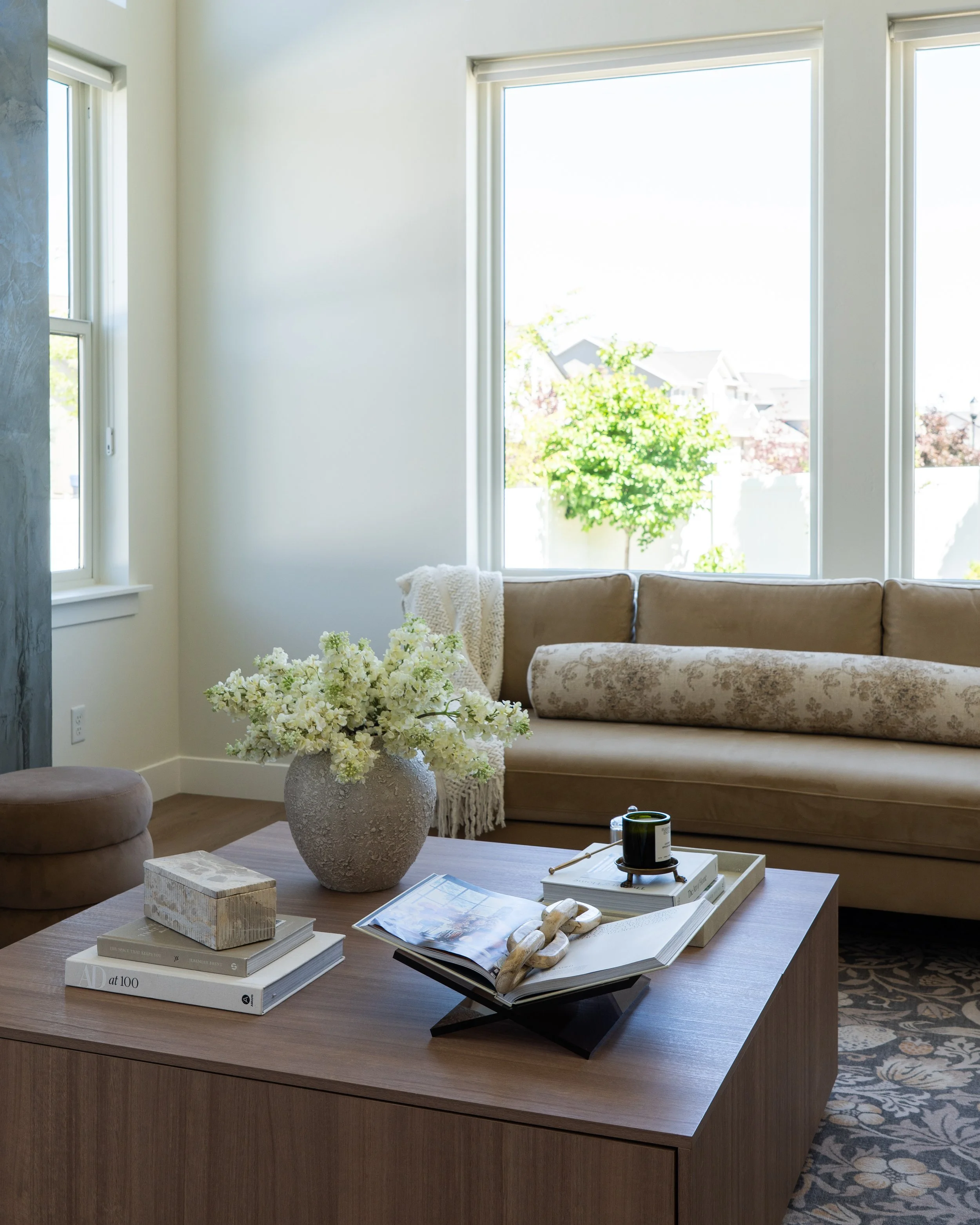 Living room corner with a wooden table holding decorative books, a vase of white flowers, and other items, a beige tufted sofa, and large windows with white frames showing a sunny outdoor view with trees.