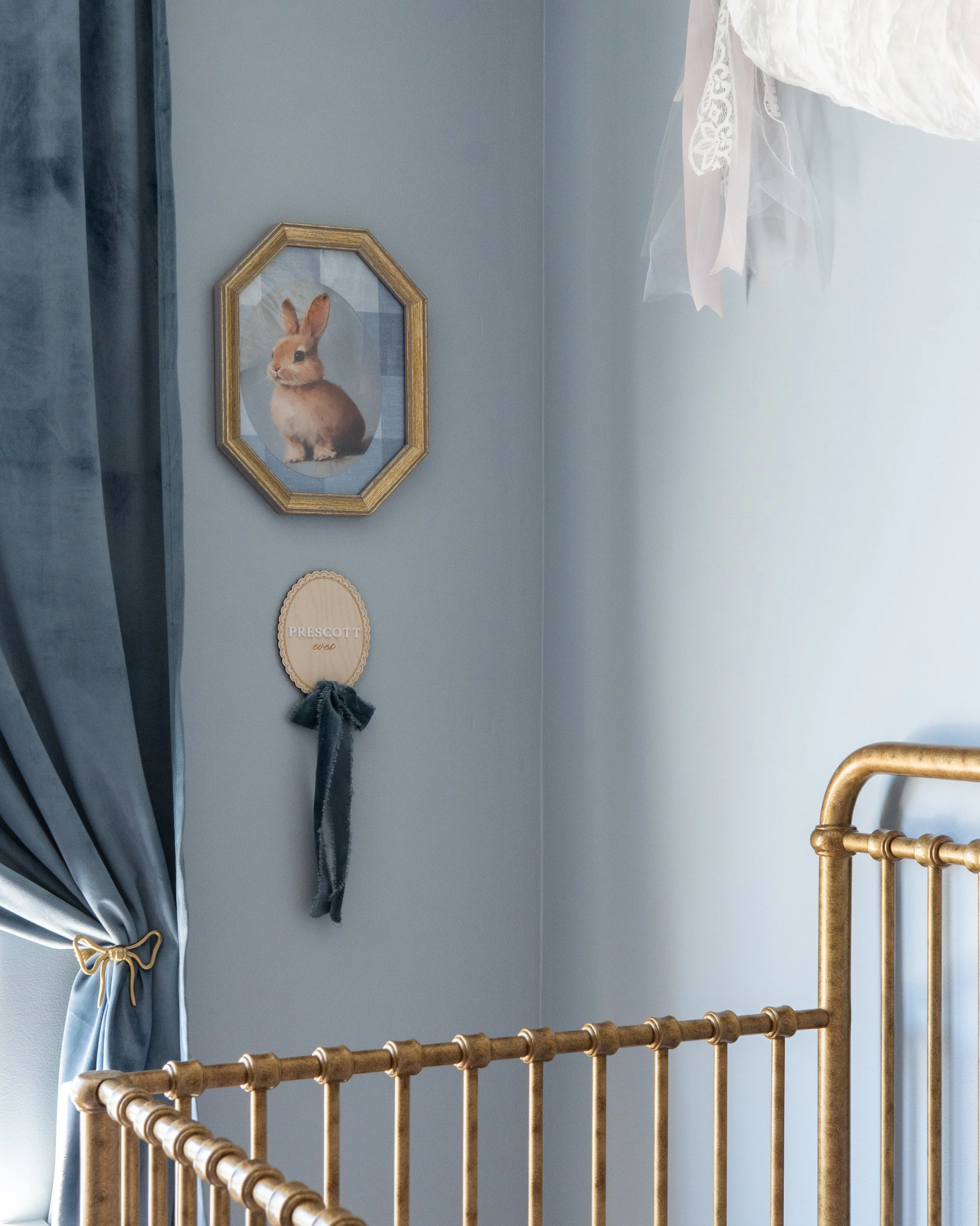 A blue wall corner with a framed picture of a rabbit, a wooden plaque with the name 'Prescott' and 'Evel,' a grey curtain with a gold bow tie, and a gold-colored metal bed rail.