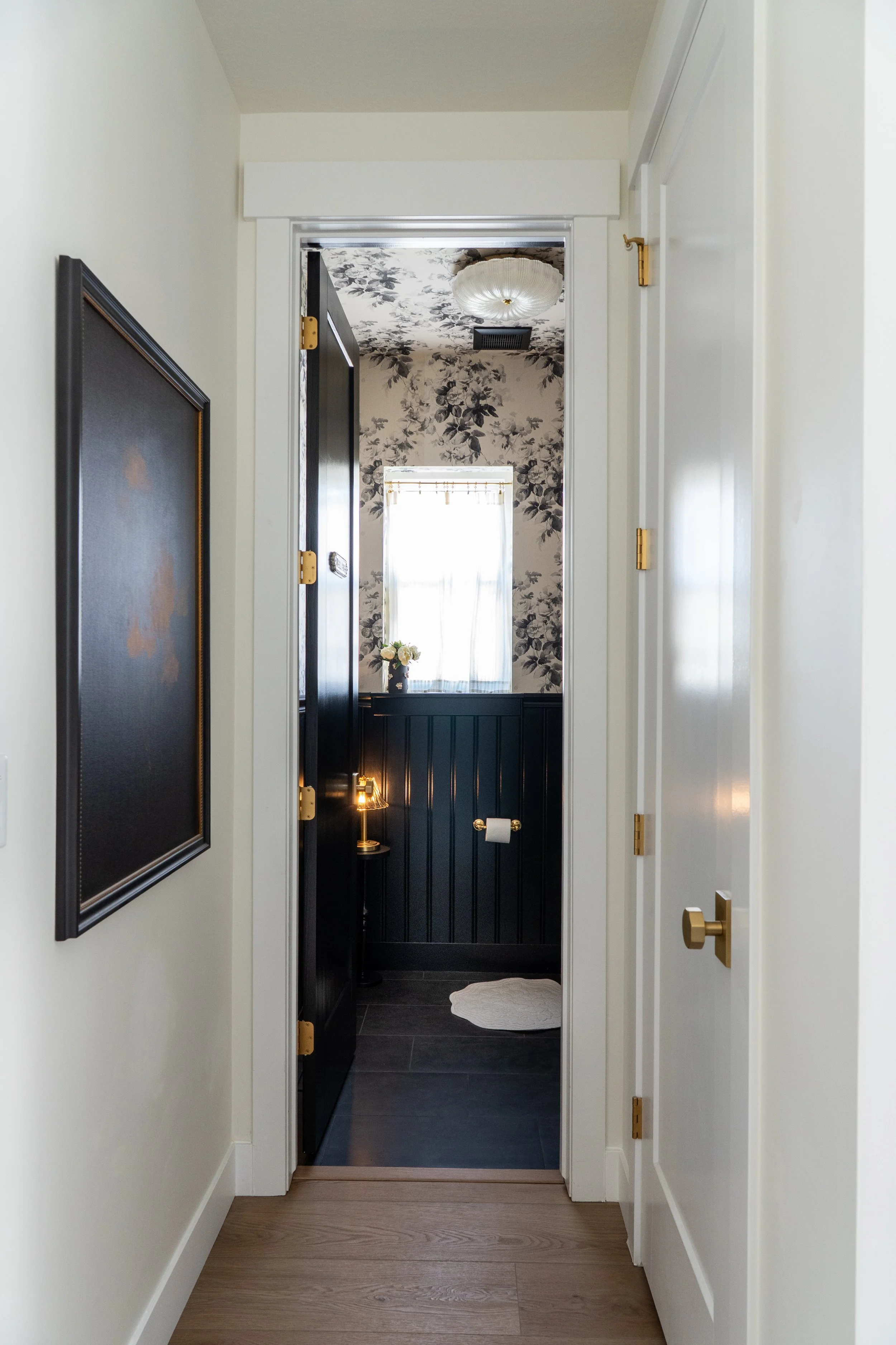 A hallway leading to a doorway with a view into a room featuring floral wallpaper, a window with sheer curtains, a small side table, a flower vase, and a ceiling light fixture.